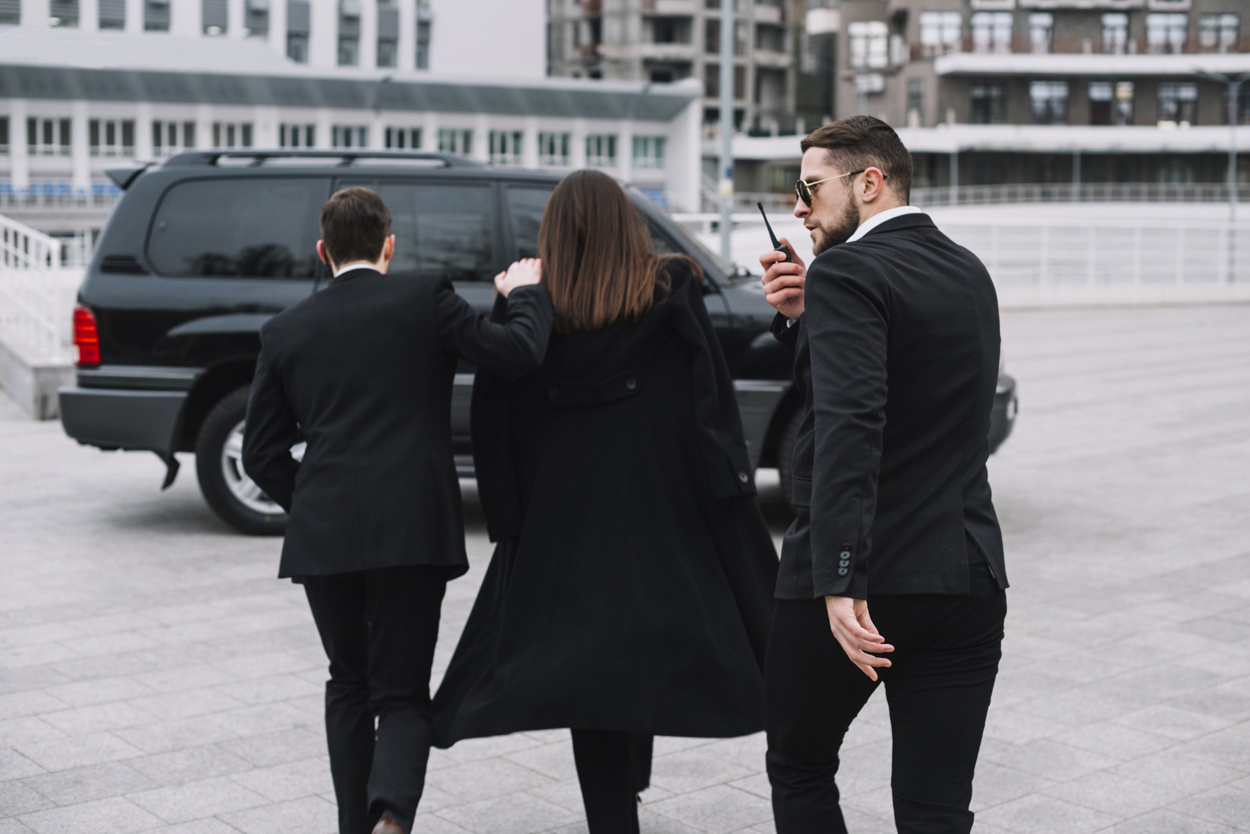 Three people in business attire walking outside near a black vehicle and urban buildings, with one man holding a walkie-talkie.