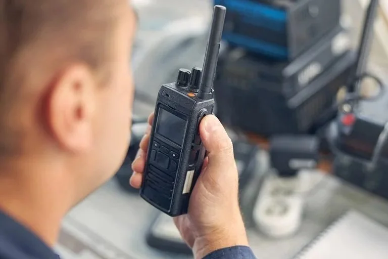Person holding a handheld two-way radio or walkie-talkie, with communication equipment and a notepad in the background.