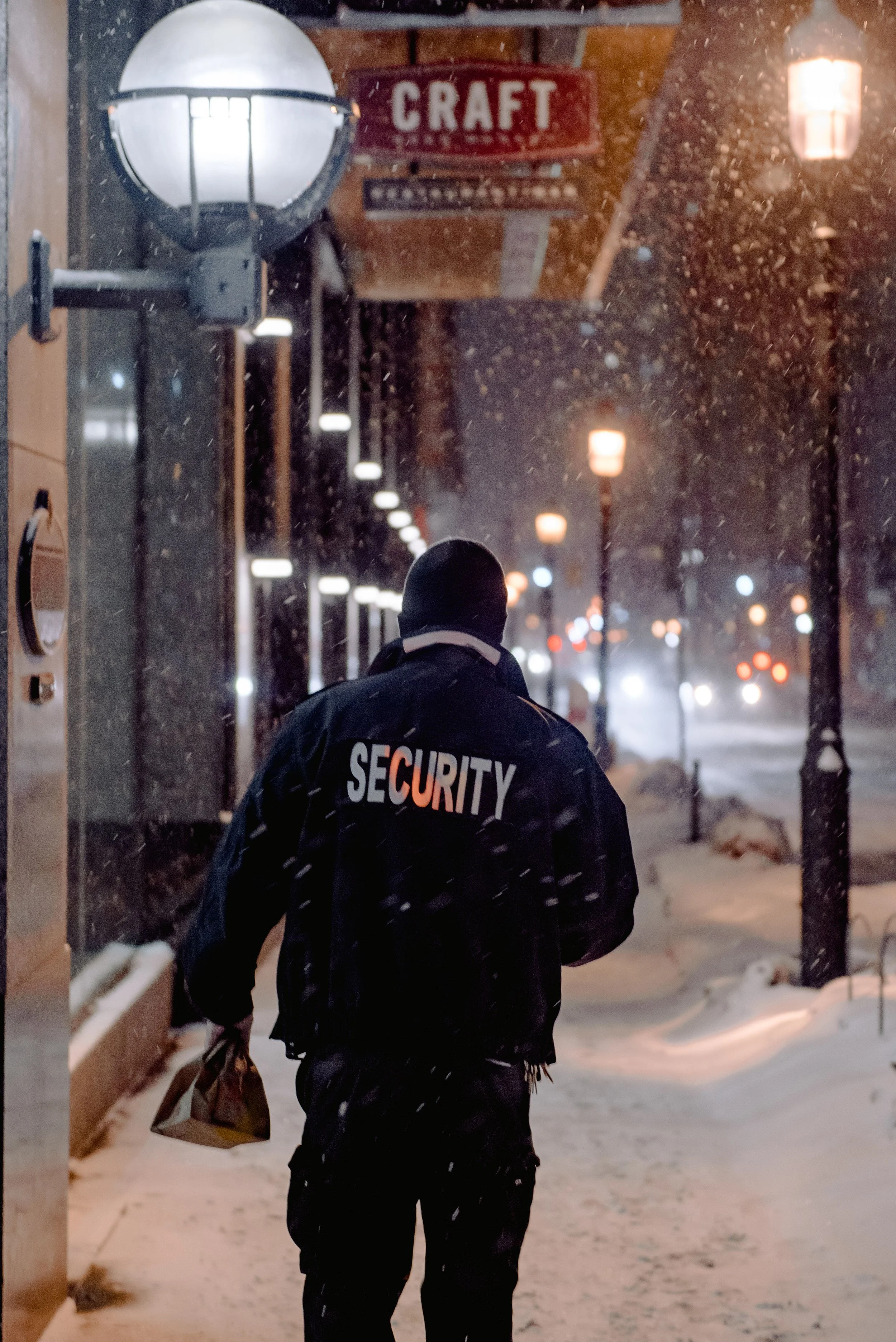 A security guard walking outdoors on a snowy night, carrying a brown paper bag, with snow falling and streetlights in the background.