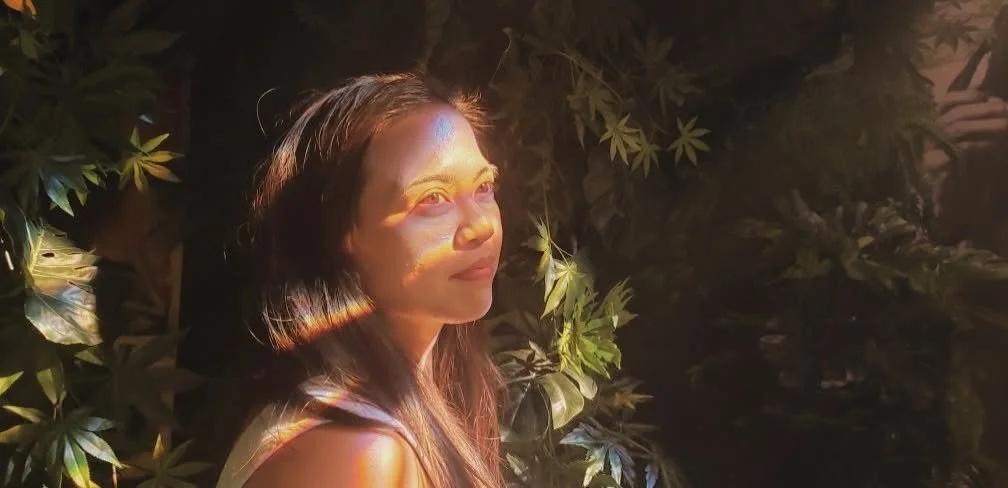 A woman with long hair faces left, surrounded by green leaves. Sunlight creates colorful light refractions on her face and shoulder.