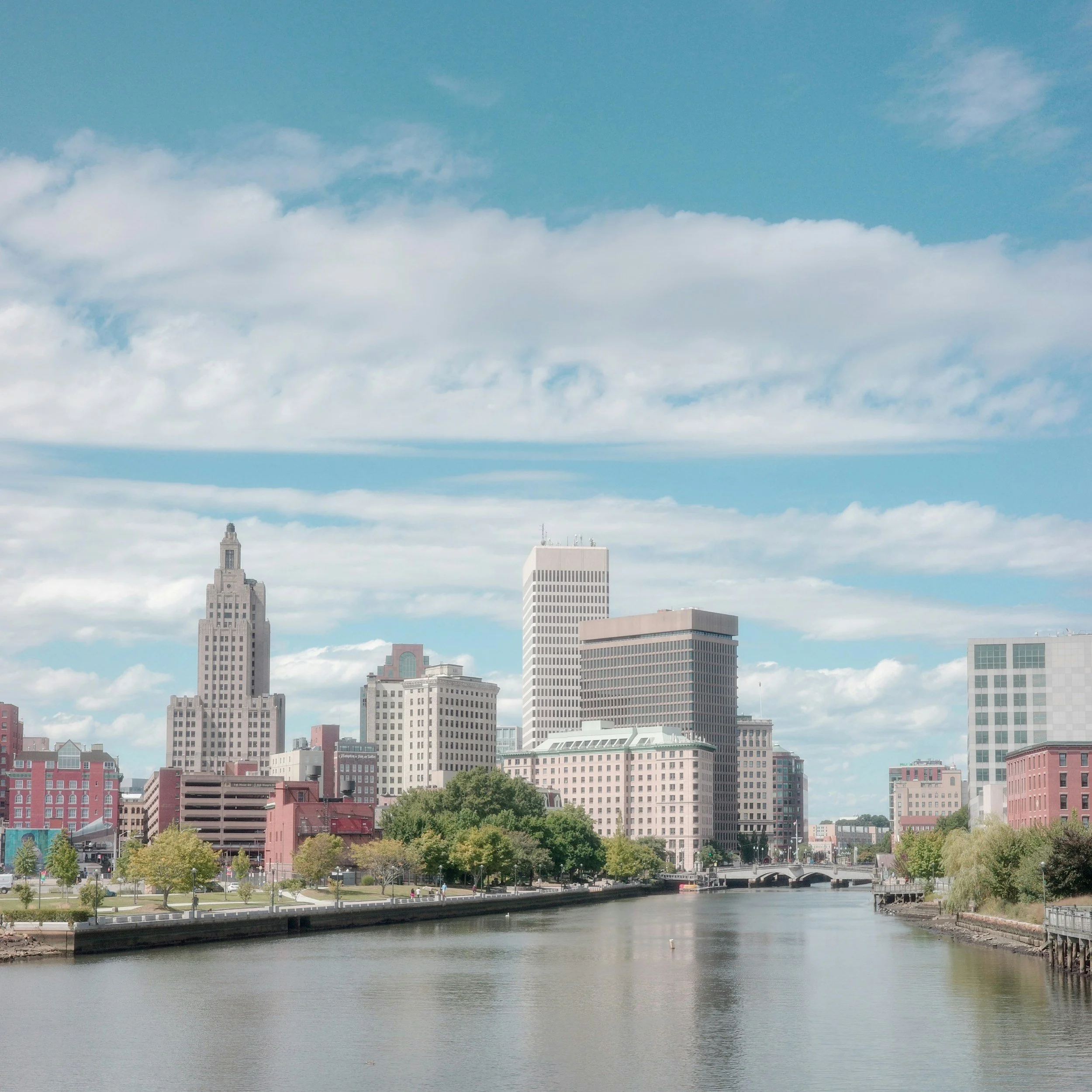 City skyline with tall buildings, a river in the foreground, blue sky, and scattered clouds.