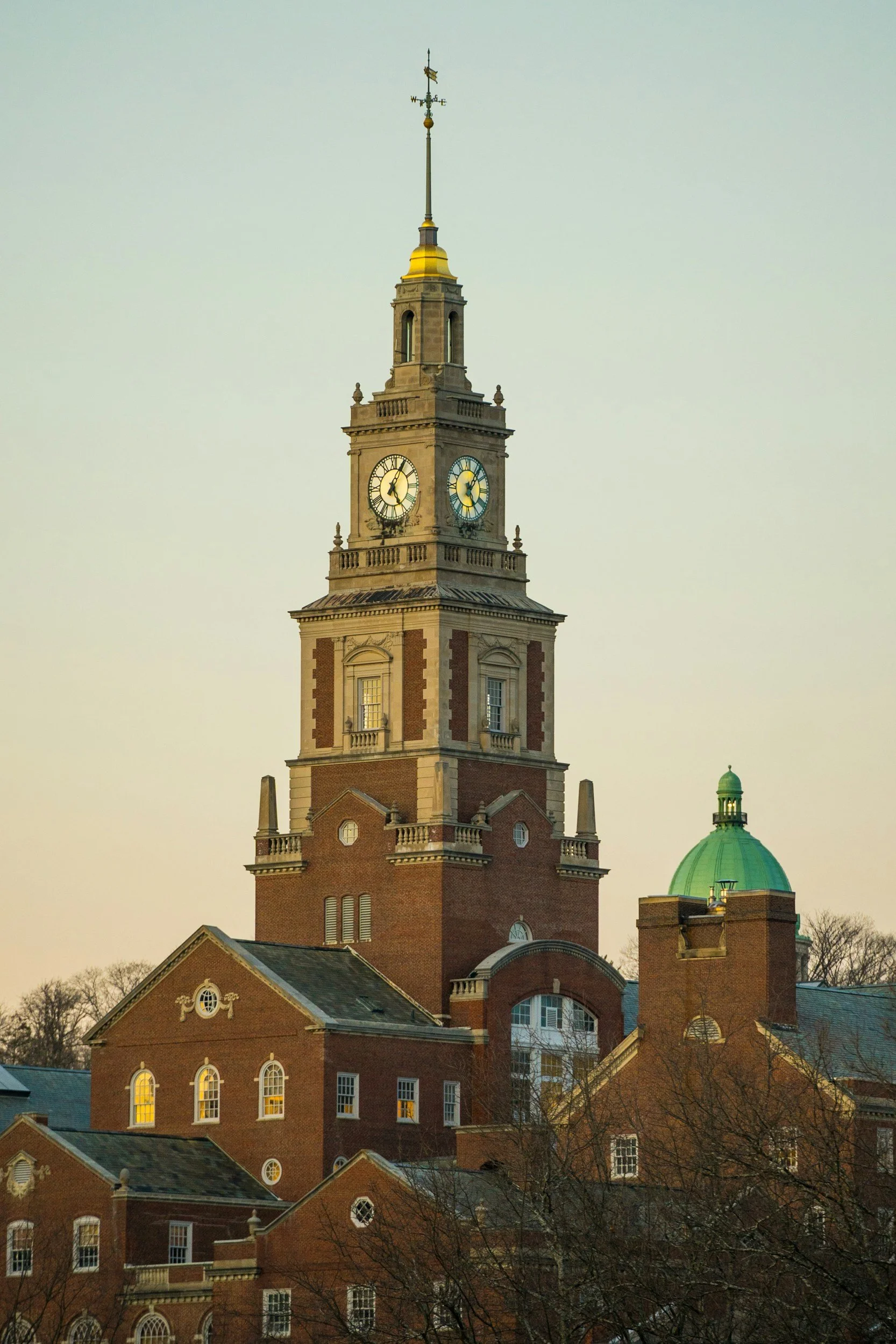 A historic brick building with a tall clock tower topped with a spire and weather vane, and a green-domed roof, against a pale sky.