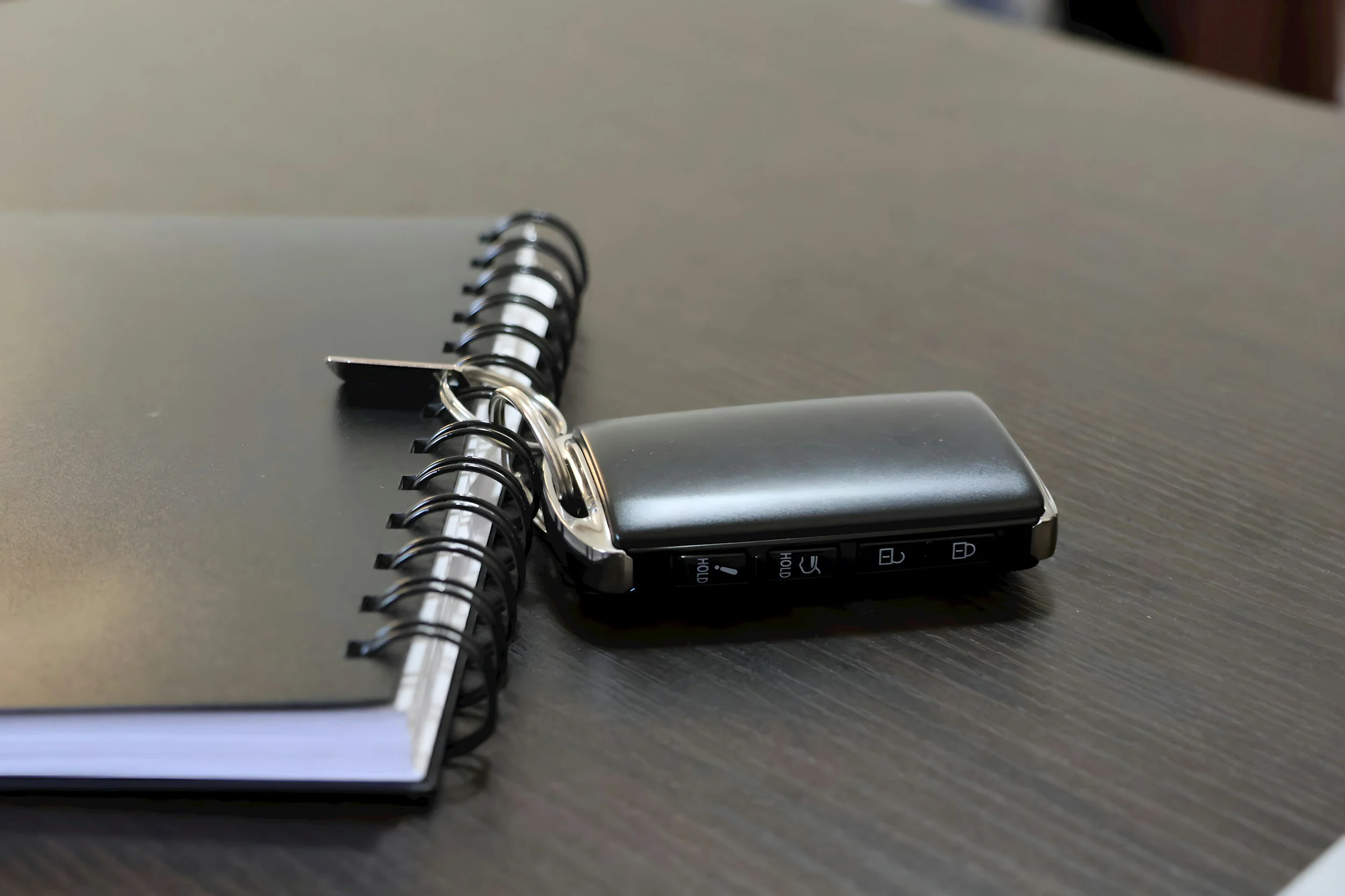 Close-up of a black spiral notebook with a metal binder clip and a car key fob resting on top, on a dark wooden surface.