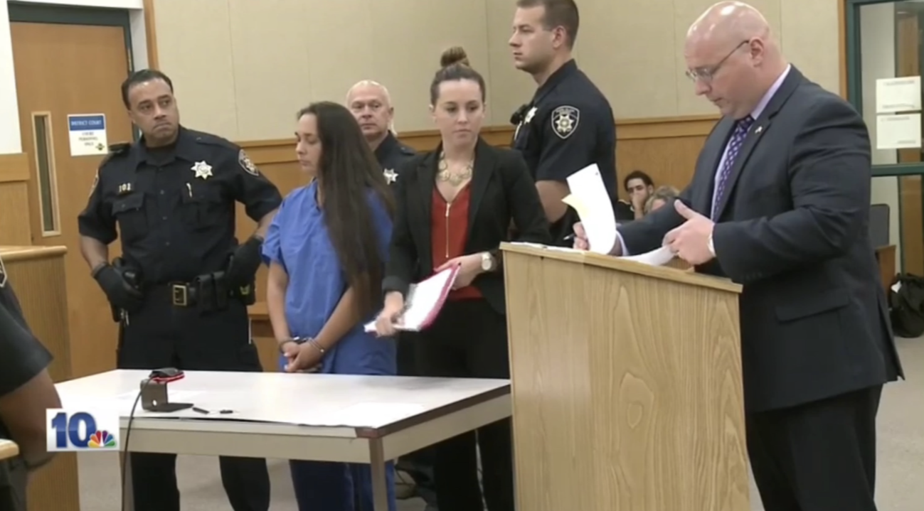 A courtroom scene with a young woman in blue scrubs standing with her hands clasped in front of her, surrounded by police officers, a woman in black, a man at a podium, and other people seated in the background.