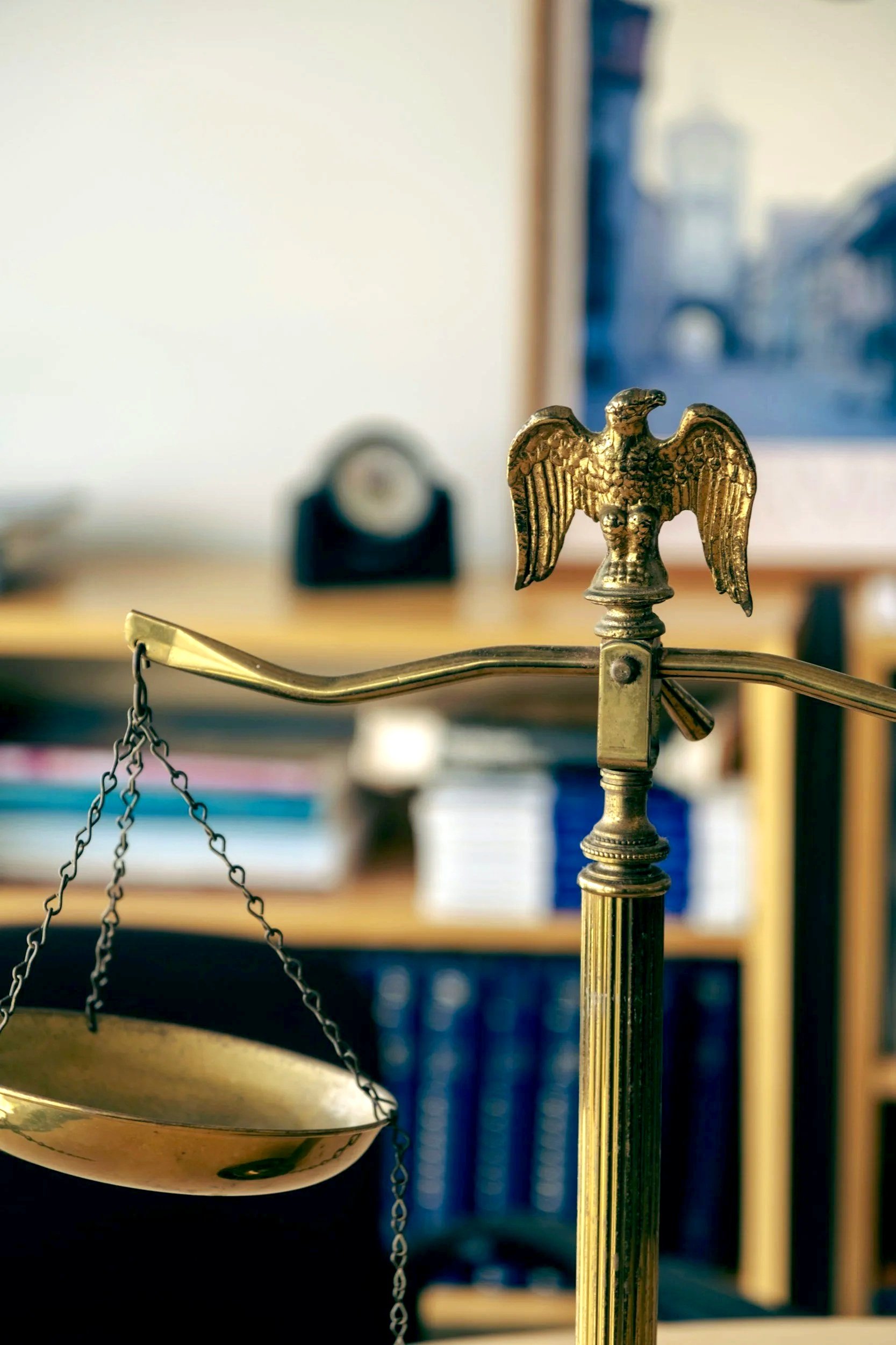 Close-up of a decorative brass eagle on a balance scale, with blurred background of books and wall.