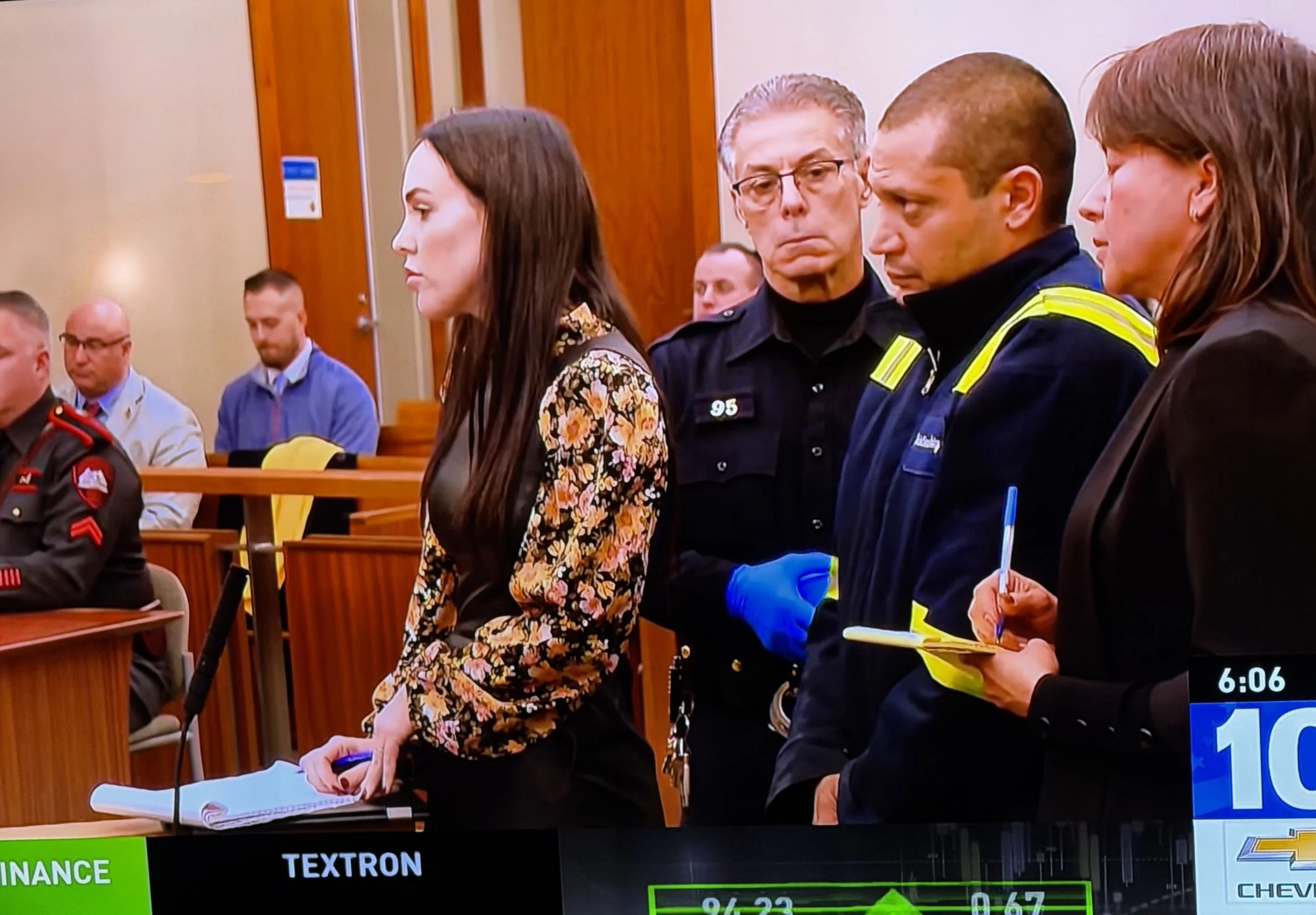 People standing in a courtroom during a legal proceeding, including a woman with long dark hair, men in police uniforms, and others sitting at benches.