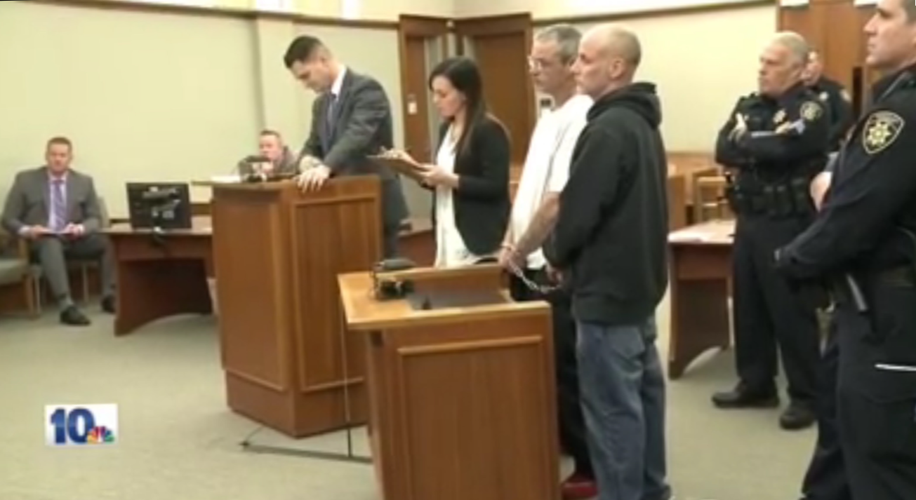 A courtroom scene with three people standing in front, including an inmate with hands restrained, and five people seated or standing in the background, including police officers.