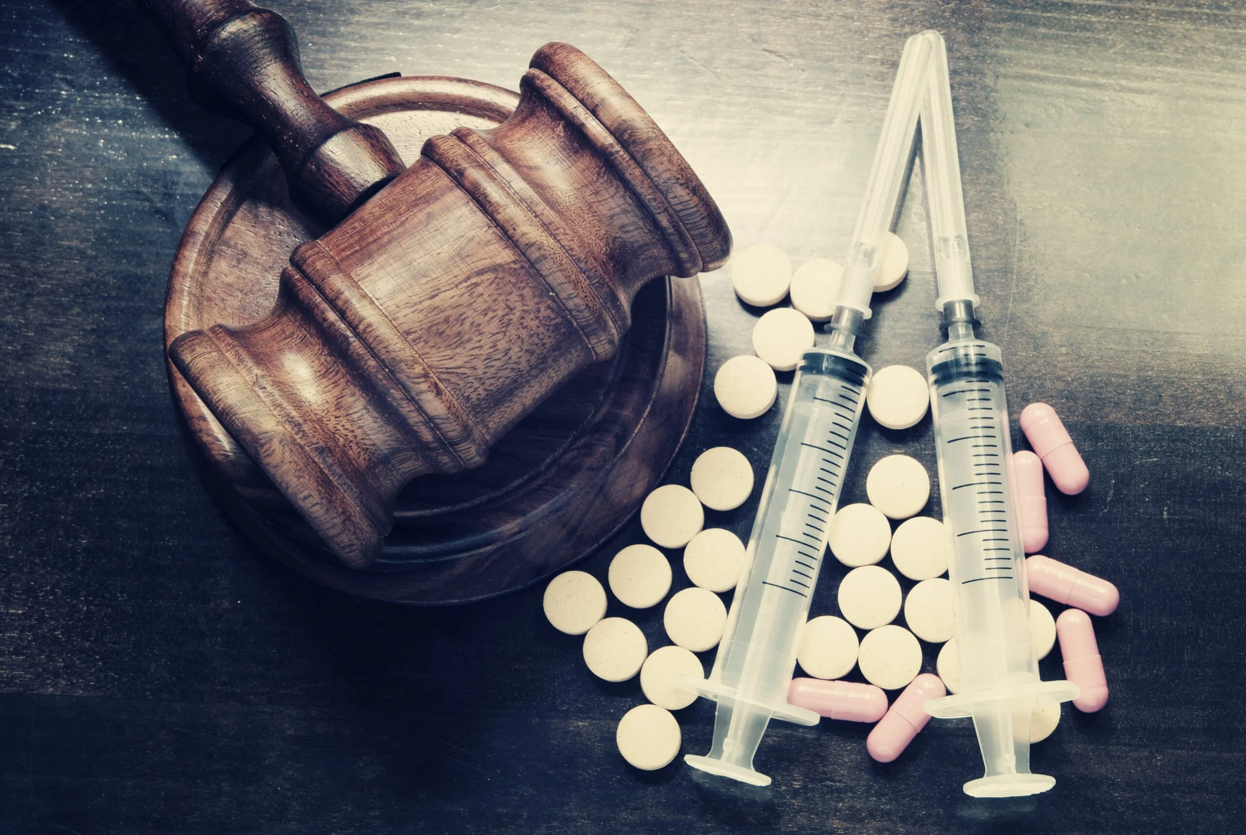 A wooden gavel lying on a round base, surrounded by white pills and several syringes with clear liquids on a dark surface.