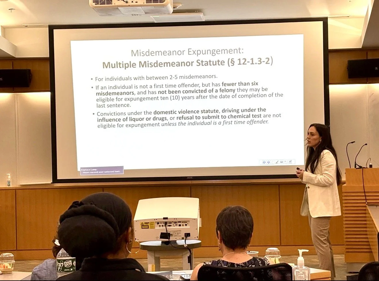 A woman giving a presentation on a large screen in a conference room. The slide discusses the Misdemeanor Expungement law, with bullet points on eligibility criteria. The audience is seated, and there are drinks on the tables.