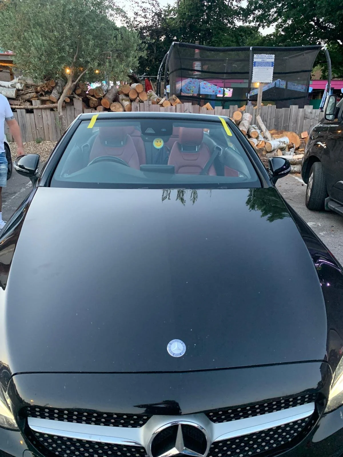 Black Mercedes-Benz car parked in a lot with logs stacked behind a wooden fence and a canopy with umbrellas above.