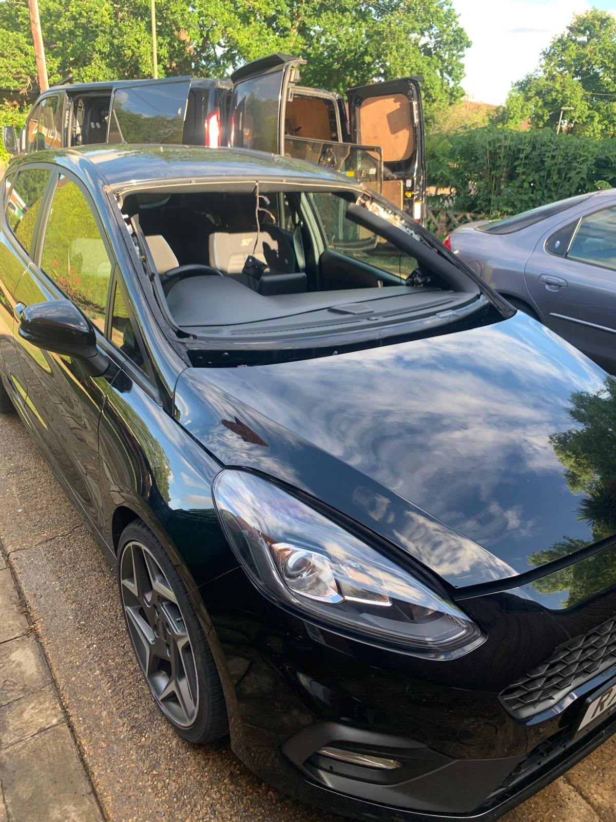 A black car parked outdoors with an open front windshield, showing a partly cloudy sky reflected on its shiny surface, and other vehicles behind it.