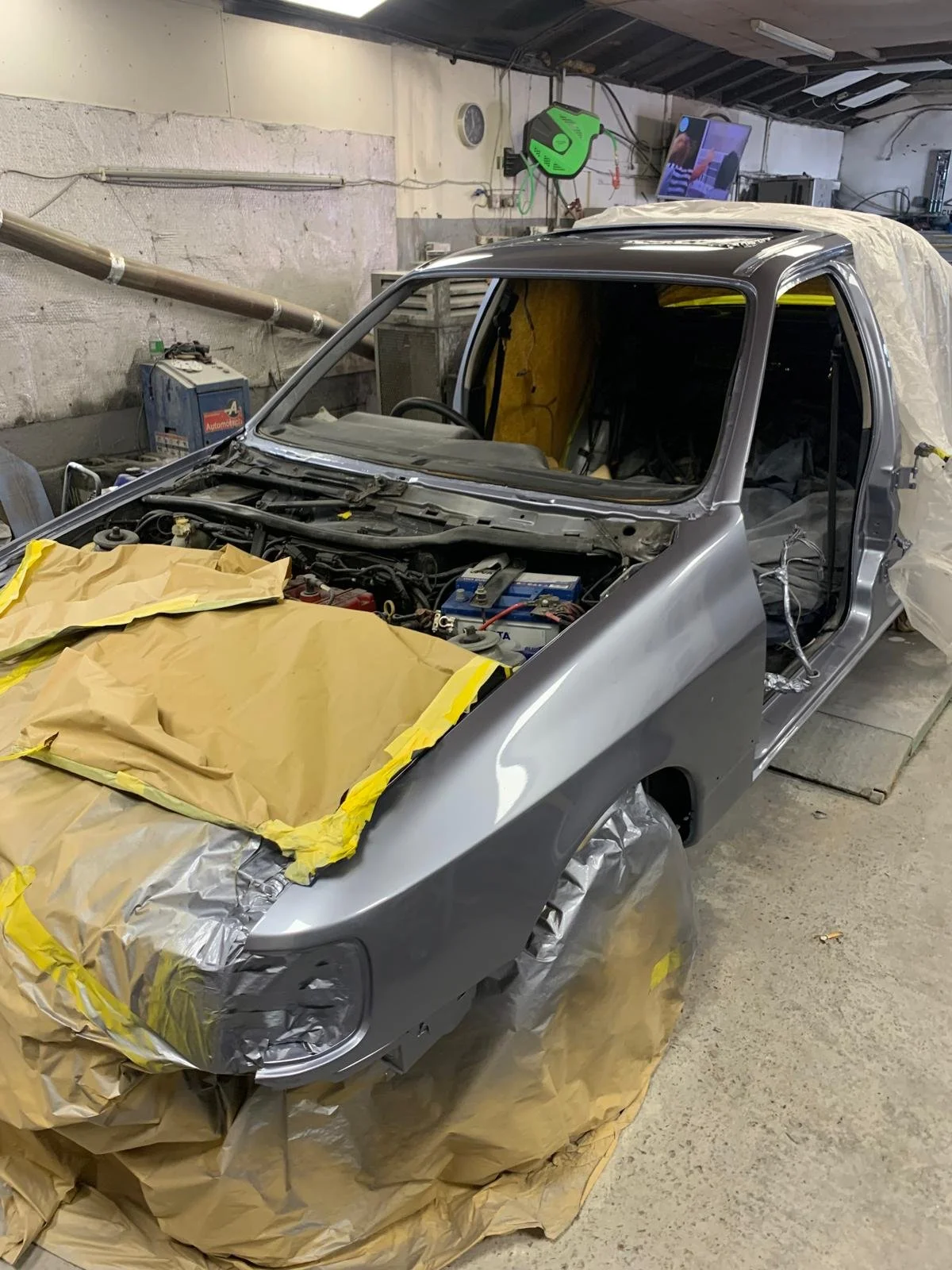 A silver car in a garage undergoing repair or painting, with painter's masking paper and tape covering sections, and work tools visible in the background.