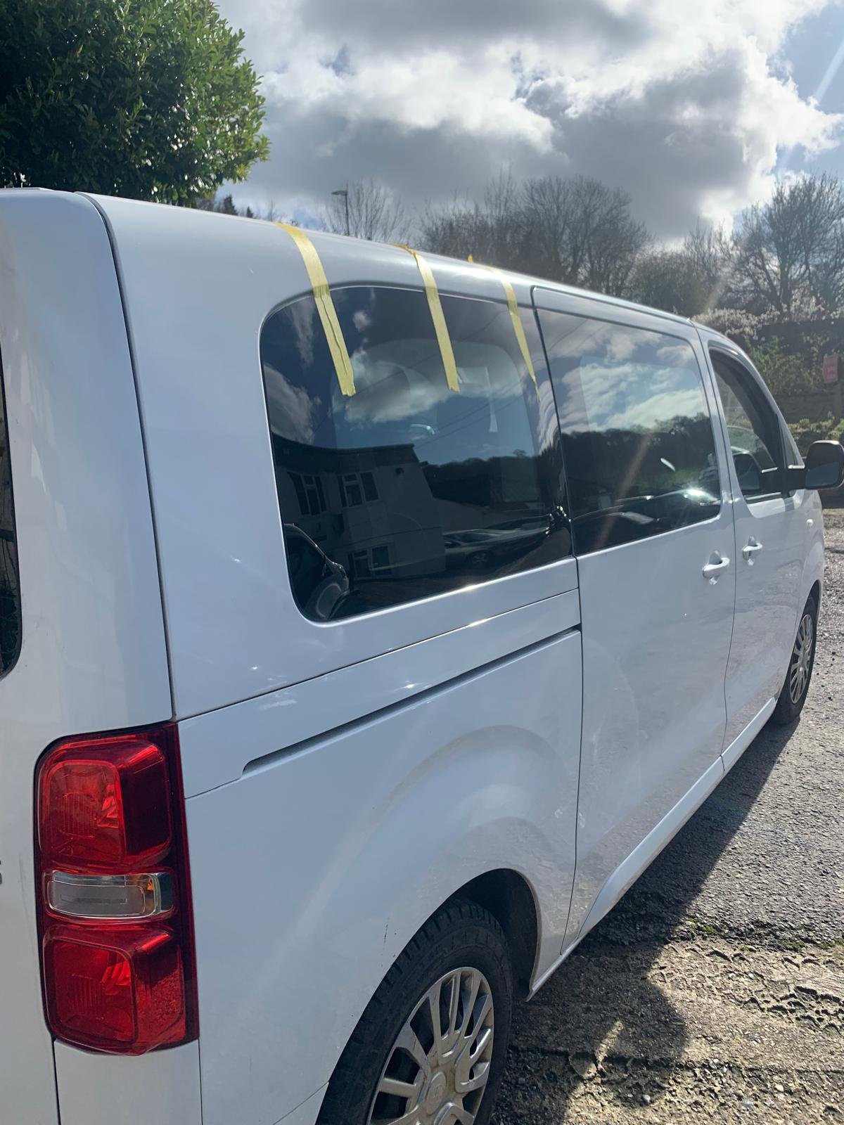 White van parked outdoors on a gravel surface with trees, cloudy sky, and sunlight reflected on the windows.