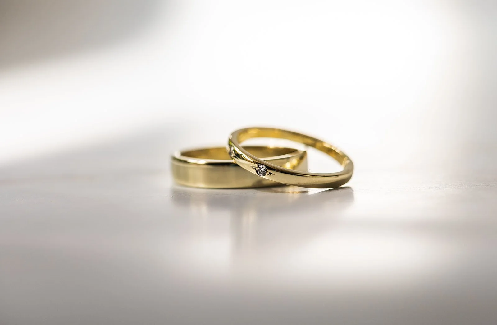 Twee gouden huwelijksringen, één met een diamant, op een lichte achtergrond.
