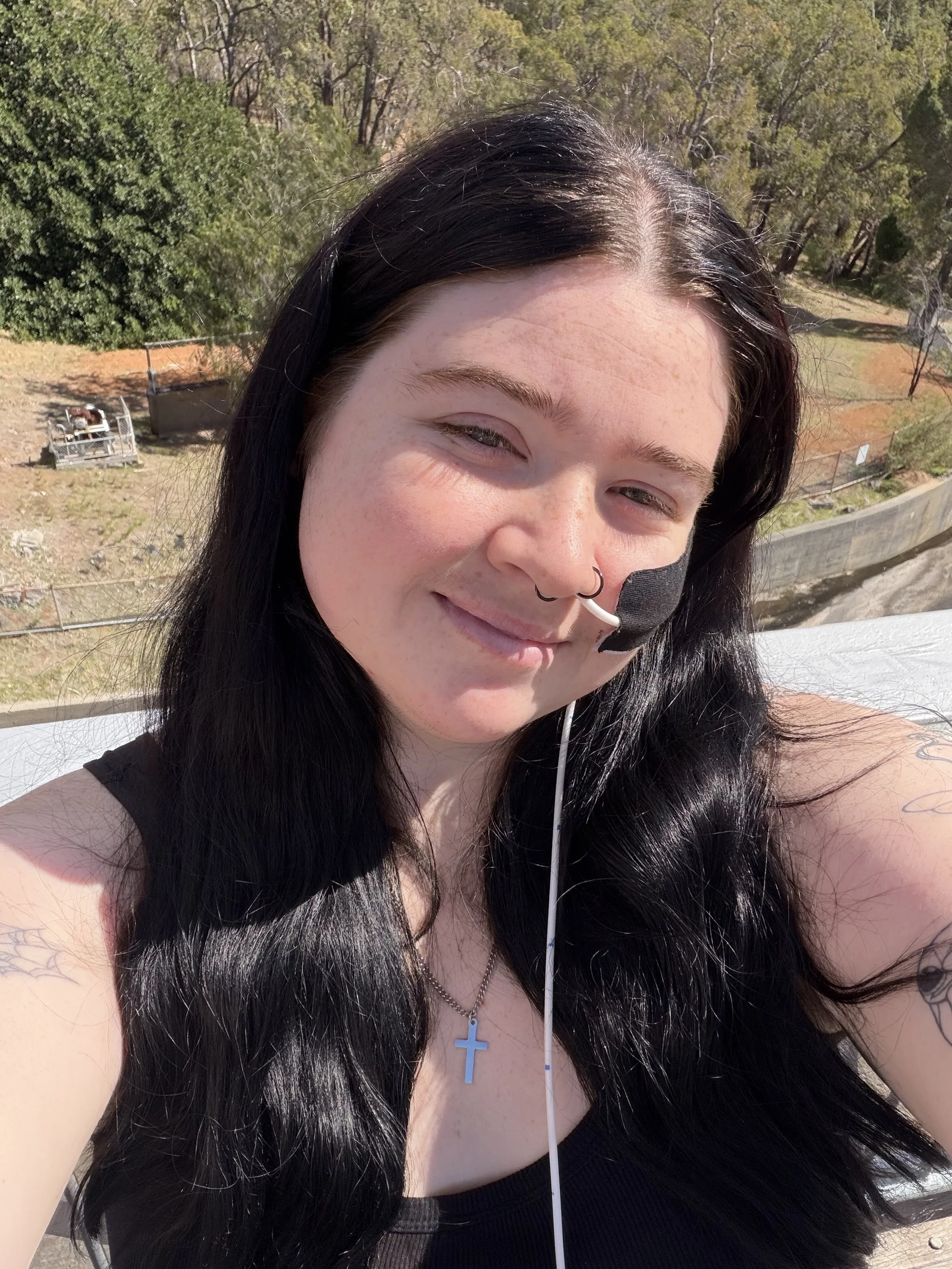 Close-up of a woman with long black hair, a septum piercing, wearing a necklace with a cross, and a black tank top, outdoors on a sunny day.