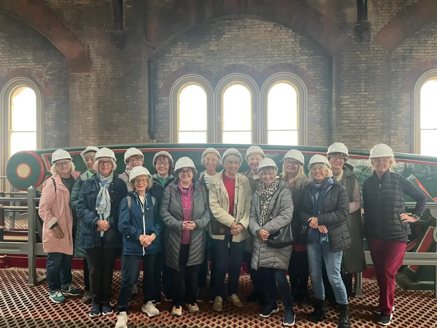 A group of elderly women and one elderly man wearing white hard hats, standing inside a building with brick walls, large arched windows, and historic industrial decor, likely on a guided tour.