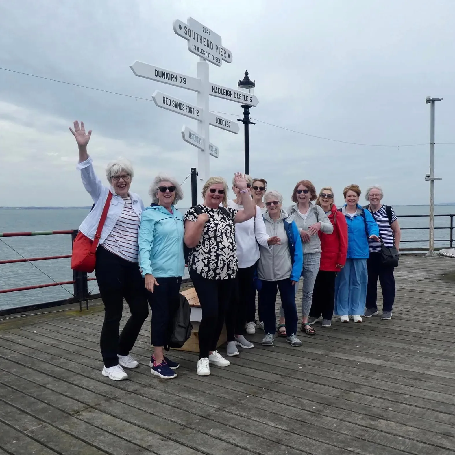 A group of smiling women standing on a pier by the water, waving, with a signpost showing directions and distances to various locations.
