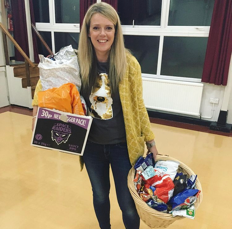 A smiling woman holding a basket filled with snacks and a box of space Raiders energy chews, standing in a room with large windows and red curtains.