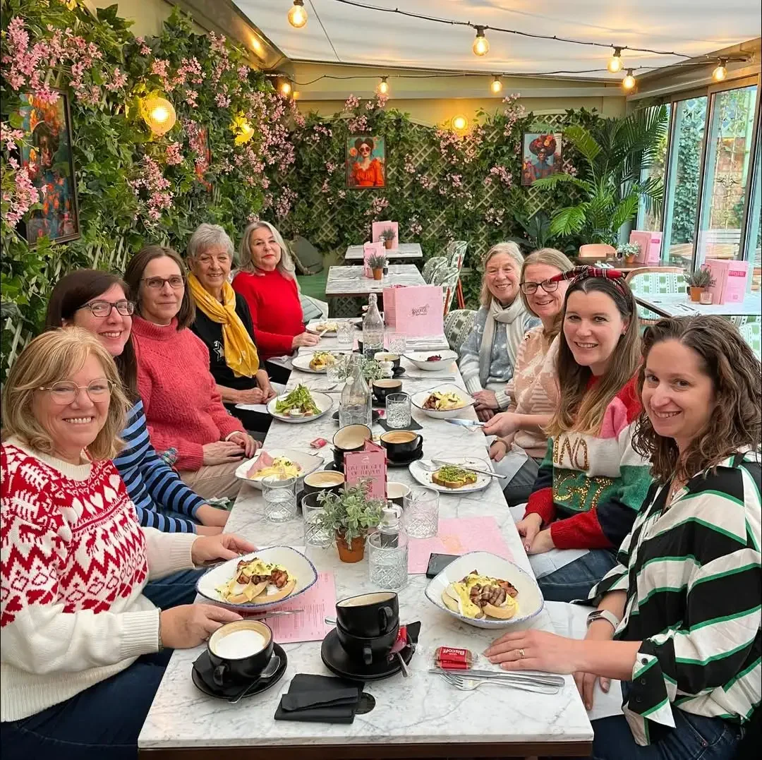 A group of women sitting around a dining table at a restaurant with pink flowers, framed portraits, hanging lights, and potted plants in the background, enjoying a meal together.