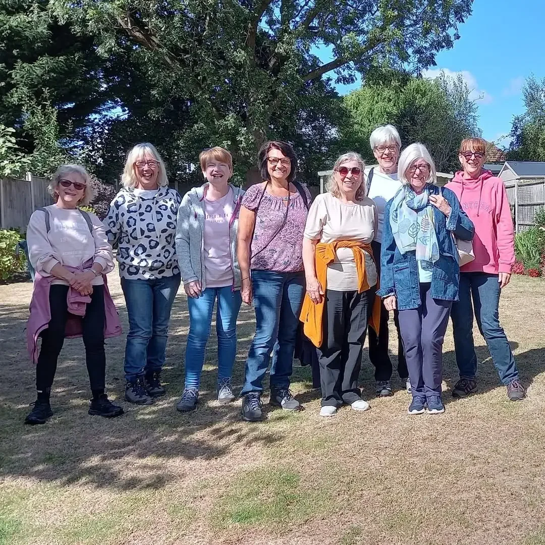 Group of nine women standing outdoors on a sunny day with trees and a fence in the background.