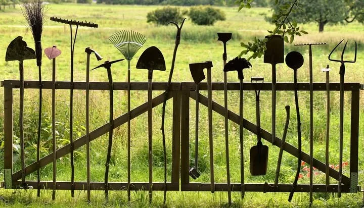 A wooden gate with various metal garden tools hanging on it, including a rake, shovel, hoe, pitchfork, and other gardening equipment, set in a green field with trees in the background.