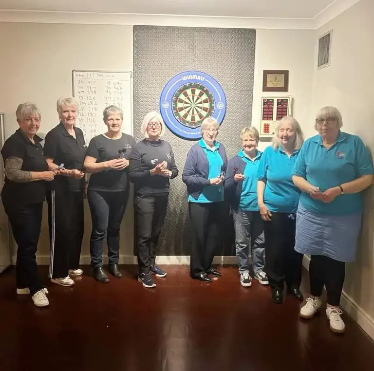 Group of seven women standing in a room with a dartboard on the wall behind them, holding darts, possibly playing darts or posing for a photo.