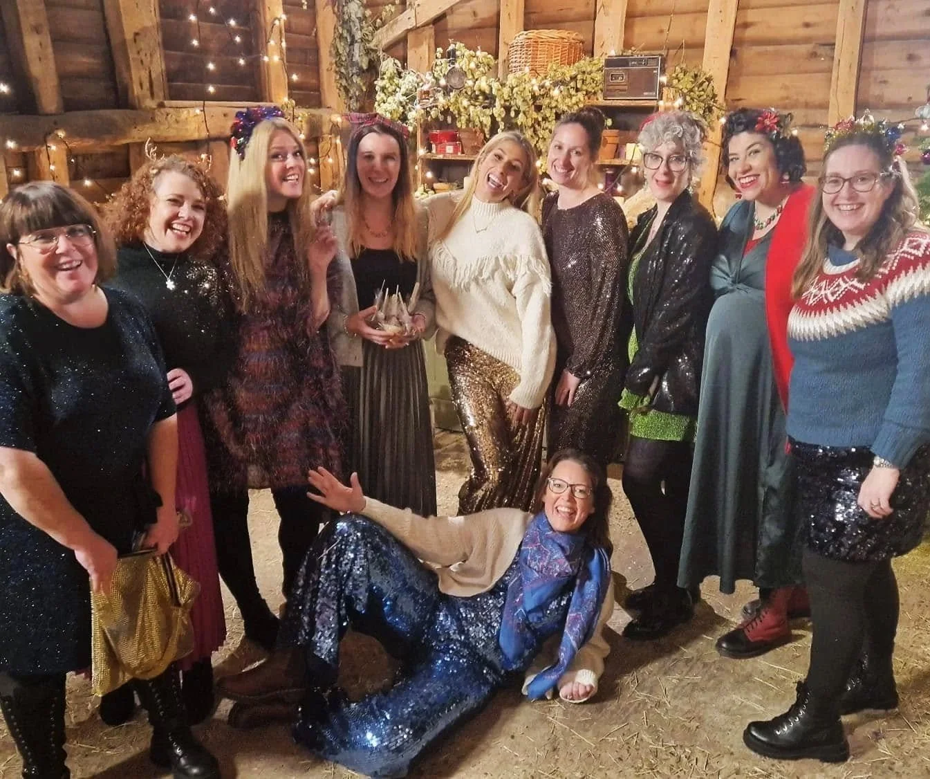 A group of women dressed festively inside a rustic wooden barn decorated with fairy lights and flowers, celebrating together.
