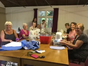 Group of women sitting around a table, sewing or doing craftwork, in a room with windows and curtains.
