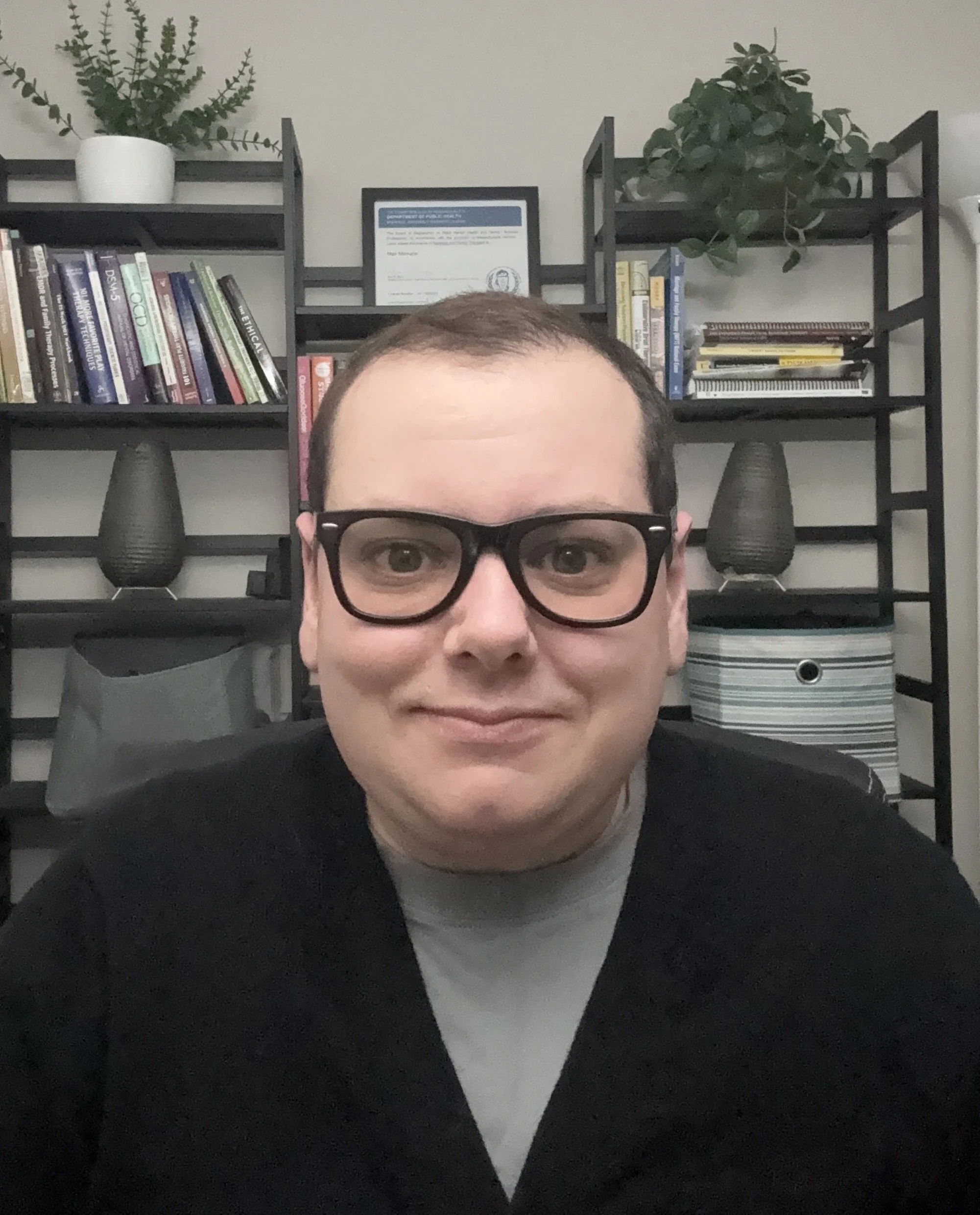 Close-up of a man wearing glasses, smiling, with a background of black shelving filled with books, plants, framed certificate, and storage boxes.