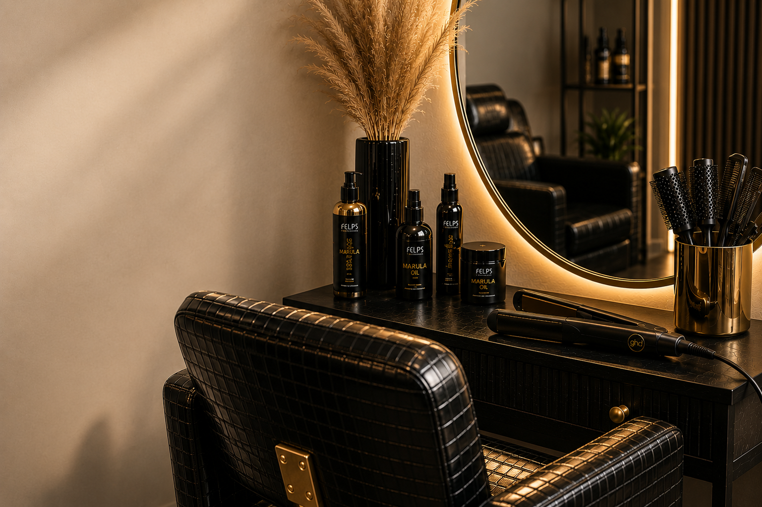 A vanity table with various hair care products, a round mirror with warm backlighting, a gold vase with pampas grass, and a container filled with black hairbrushes.