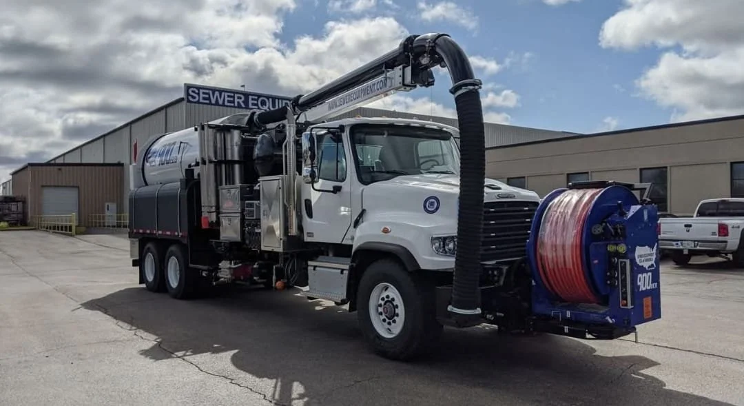 A large white utility truck equipped with a retractable tree-trimming bucket and heavy-duty hoses, parked on the side of a road with trees and a partly cloudy sky in the background.