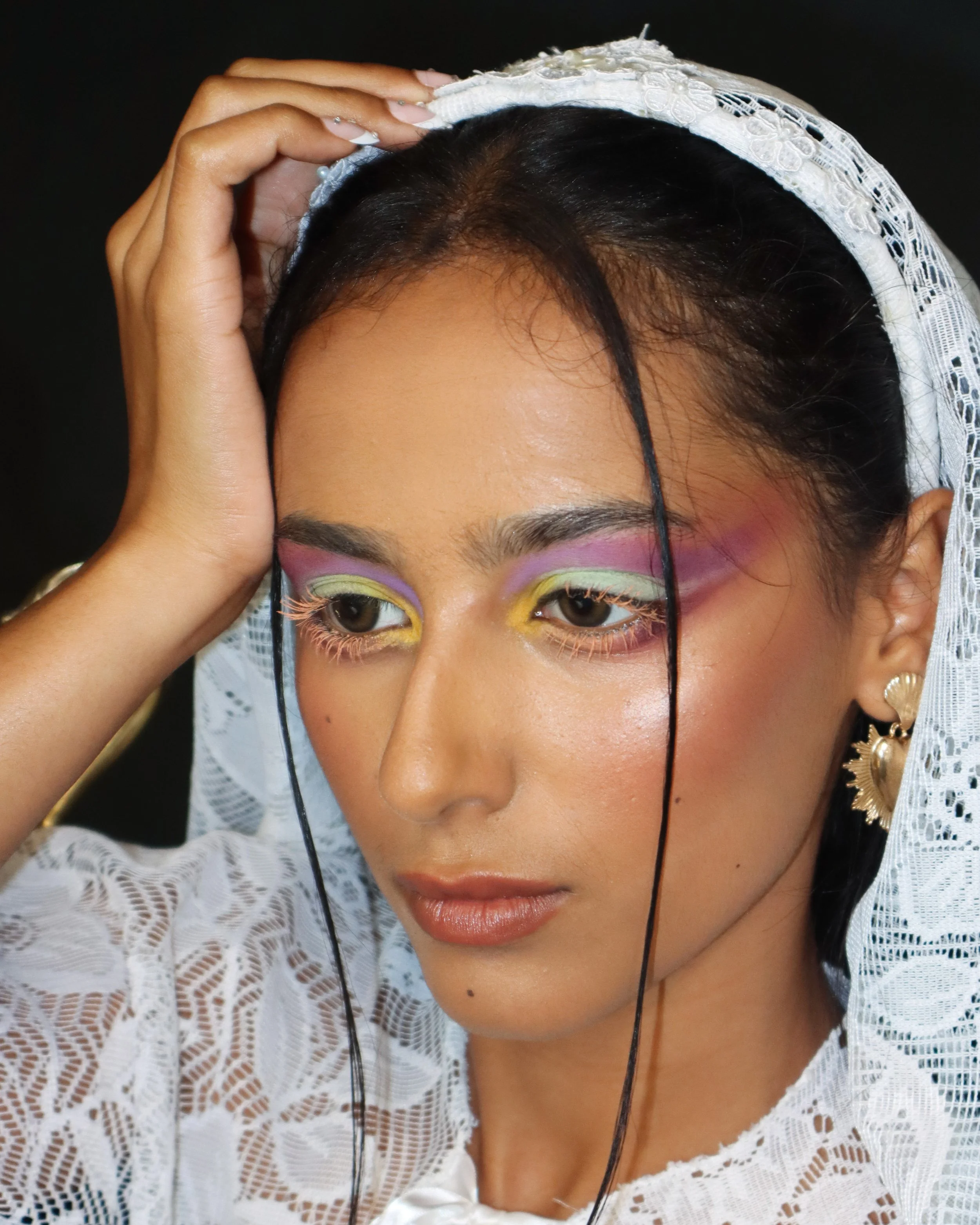 A woman with colorful makeup, wearing a white lace head covering, holding her hair back with her hand, and wearing gold and pearl earrings.