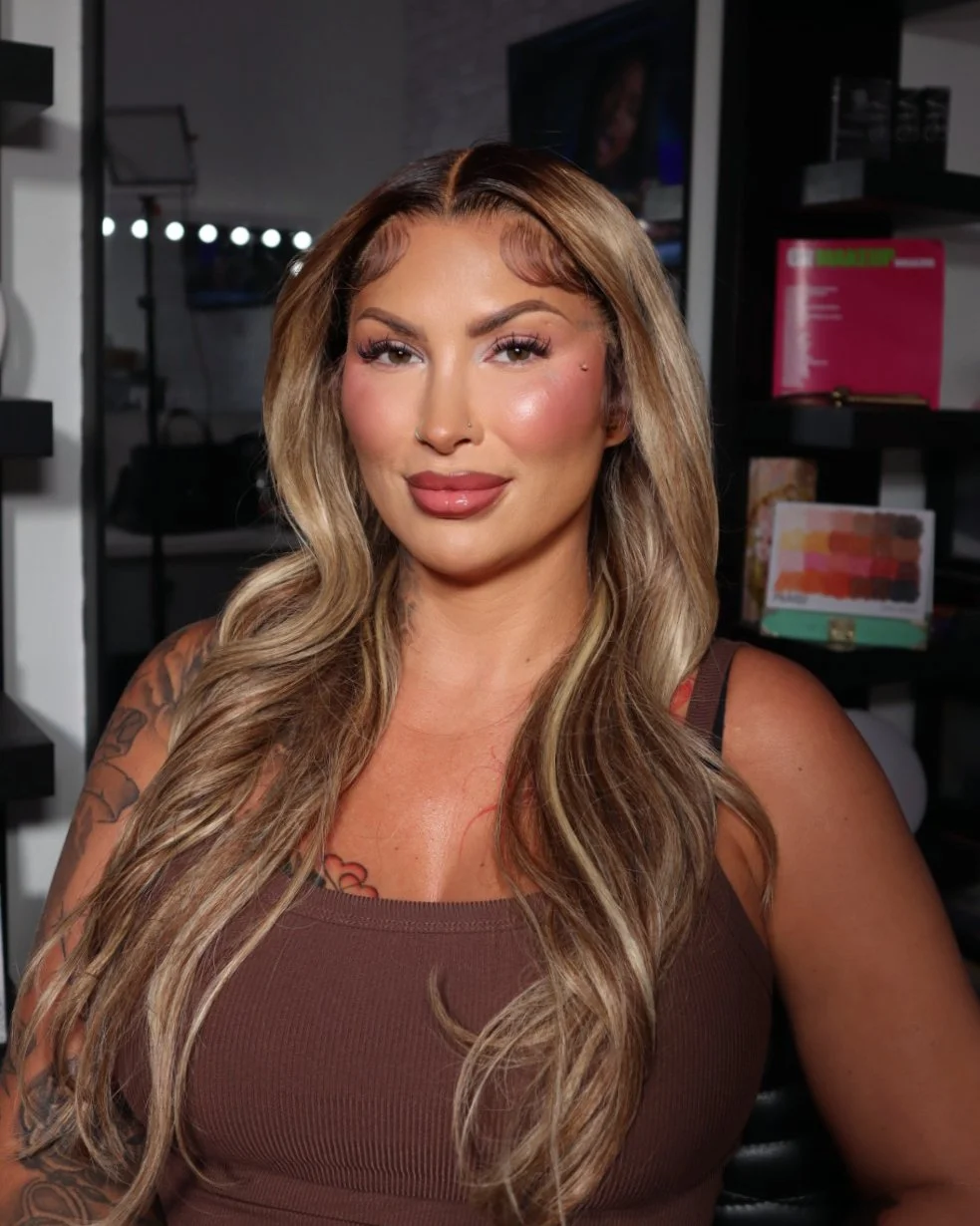 A woman with long, wavy blonde hair and facial makeup, smiling in an indoor setting with makeup products and a TV in the background.