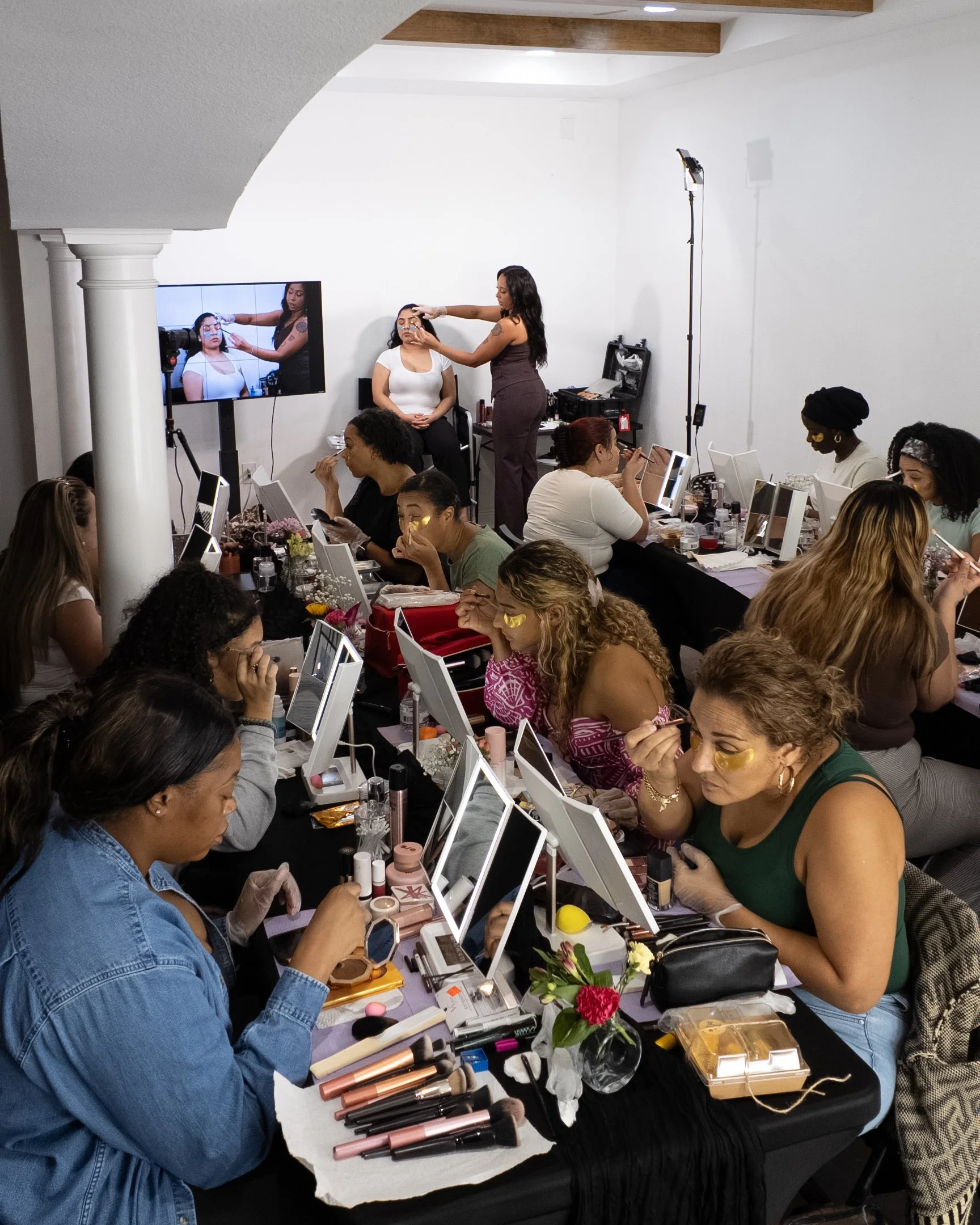 A group of women at a makeup workshop, seated at a long table covered with various makeup products and brushes, with a woman demonstrating makeup techniques. Some women are applying makeup and others observe. A large mirror with makeup tools, flowers, and makeup kits are on the table. In the background, a woman is sitting in front of a television monitor that displays a close-up of her face with makeup being applied.