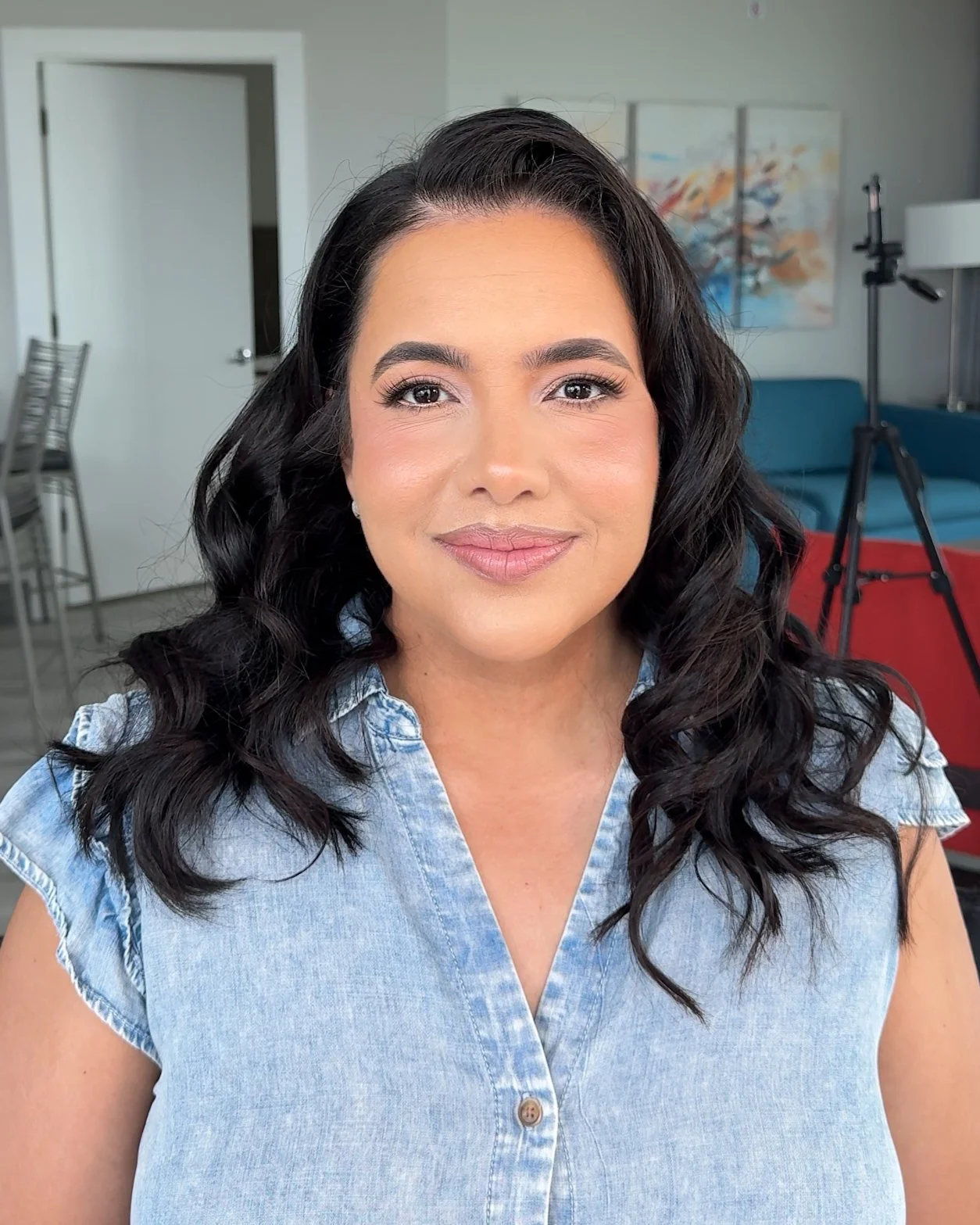 A woman with long, wavy black hair, wearing a light denim shirt, smiling in a bright indoor setting with a blue sofa, colorful artwork, and a tripod in the background.