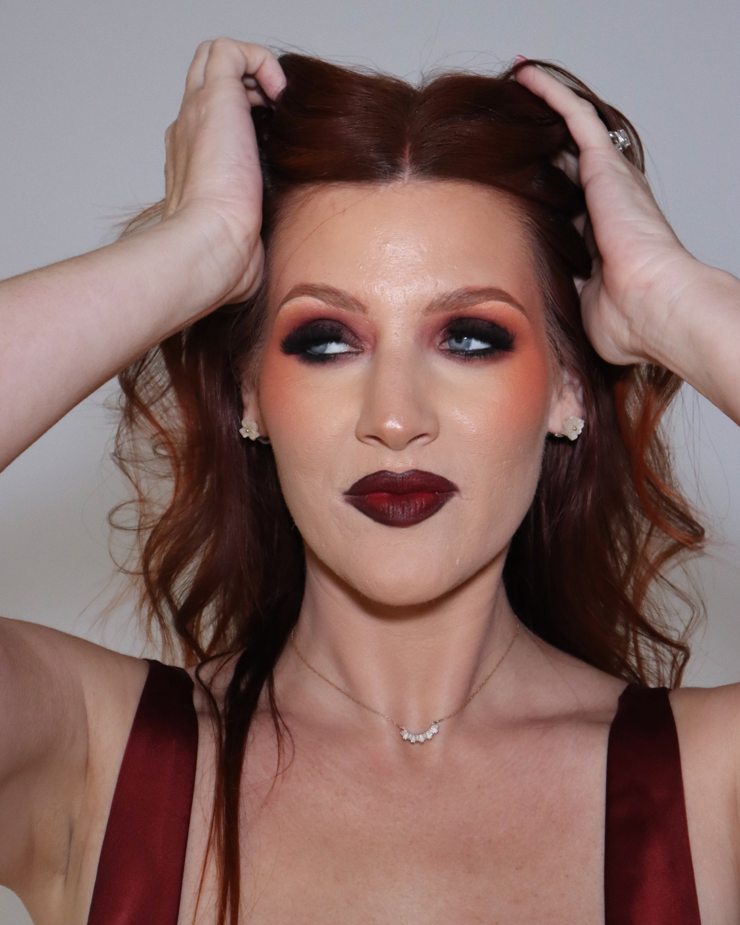 A woman with red hair, dramatic eye makeup, and dark lipstick holding her hair back with both hands, wearing a dark top and a delicate necklace.