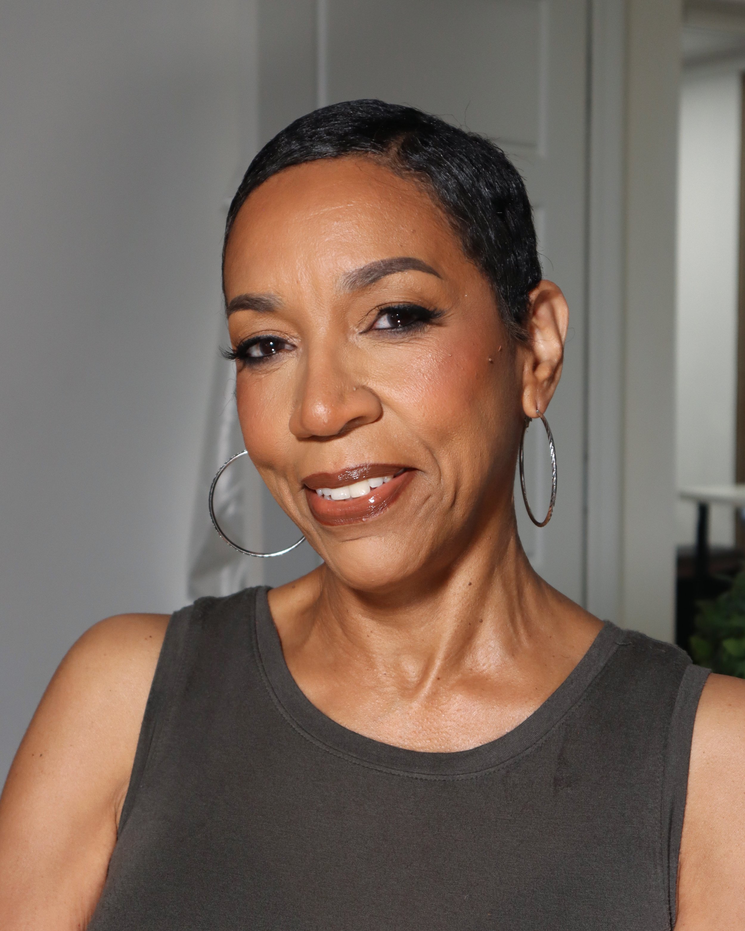 Close-up of a smiling woman with a dark sleeveless top, hoop earrings, and short black hair styled in a sleek pixie cut, standing in front of a neutral background.