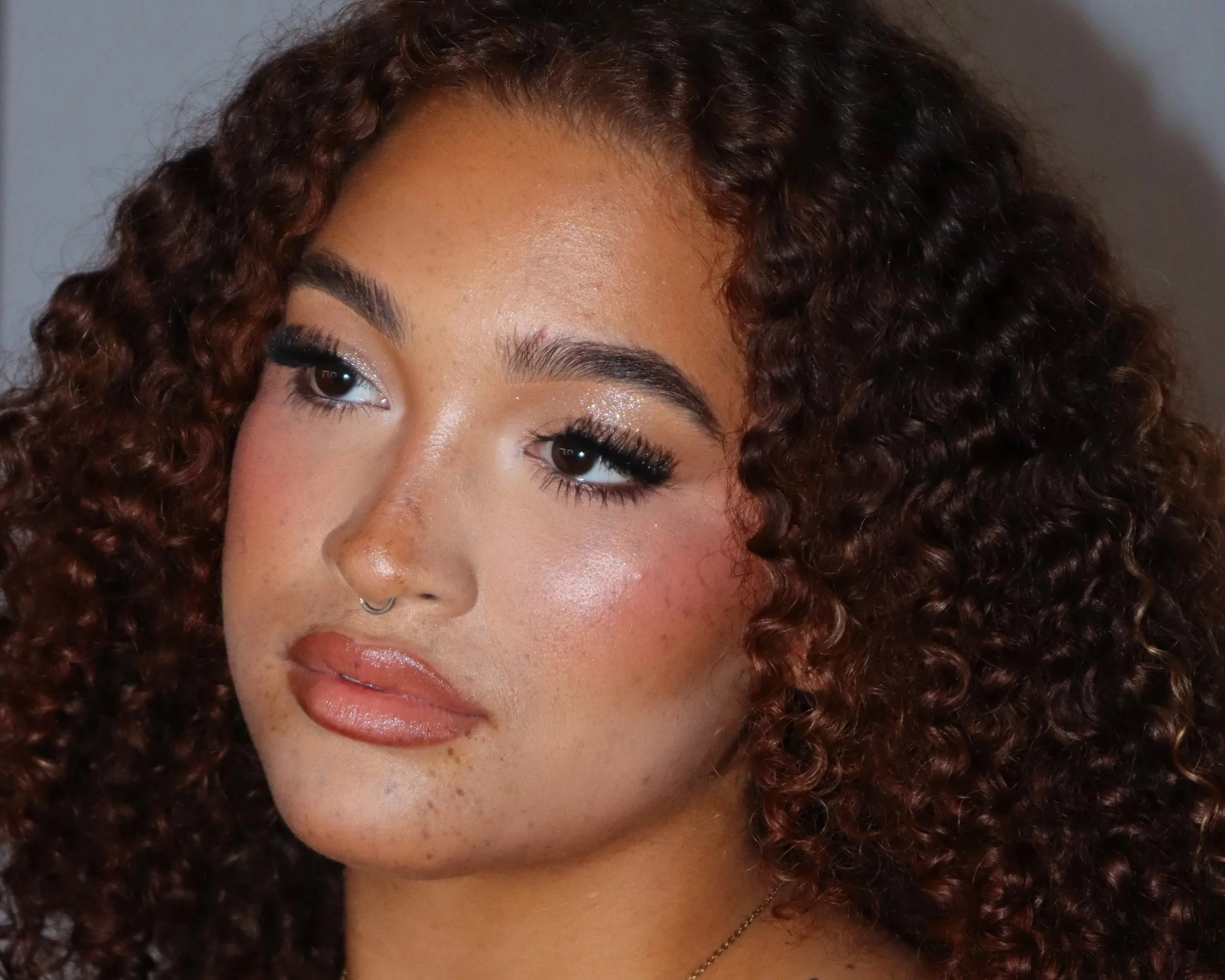 Close-up of a woman with curly, reddish-brown hair, makeup including glitter eyeshadow, long eyelashes, well-defined eyebrows, and glossy lips, wearing a thin necklace.