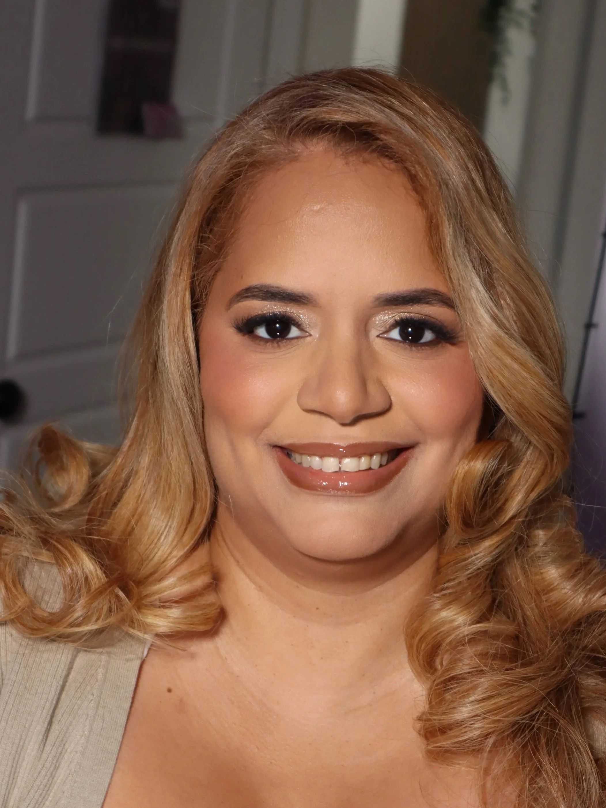 A woman with long, wavy blonde hair smiling at the camera, with makeup including eye shadow and lipstick, in a room with a dresser and mirror in the background.