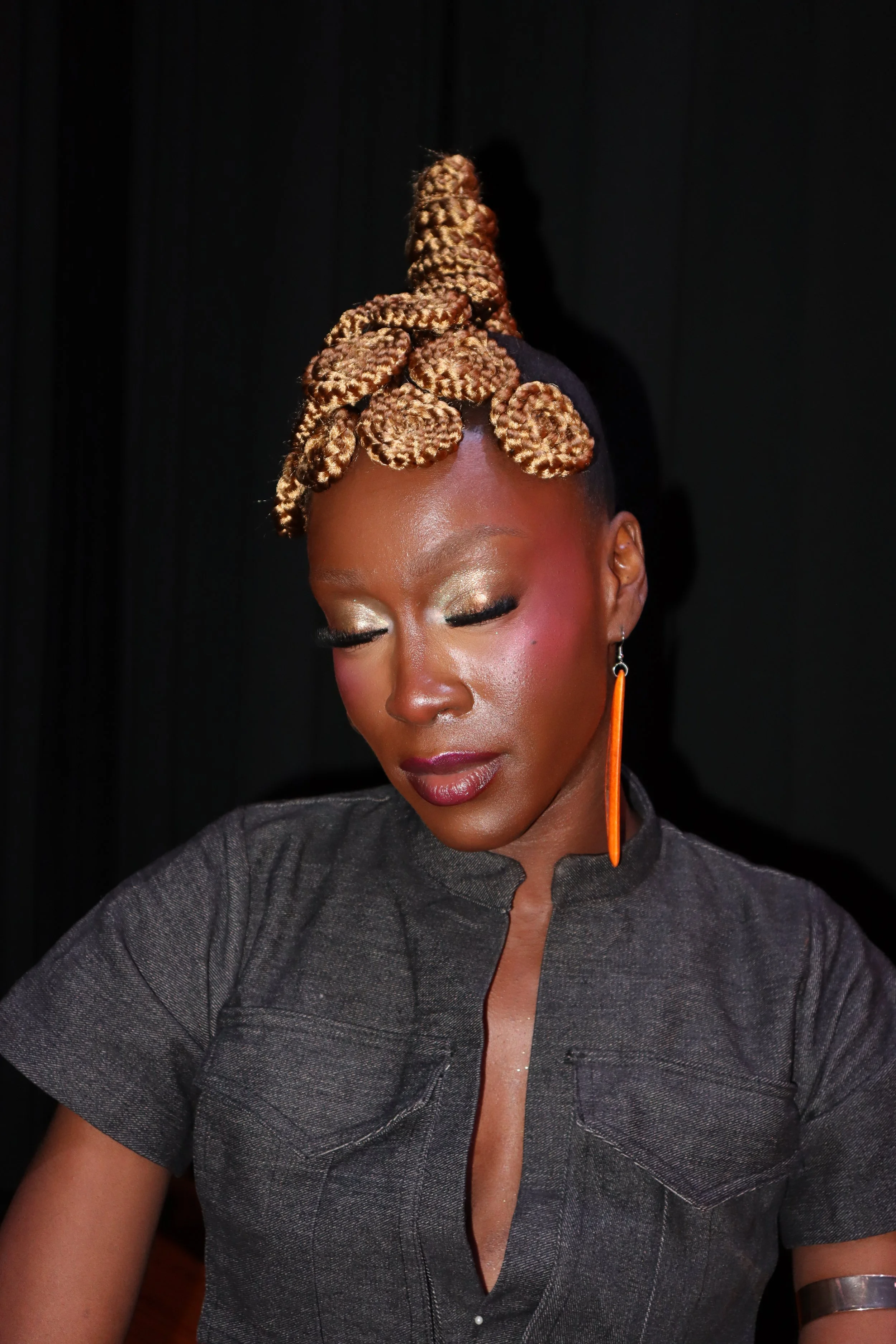 A woman with golden, tightly styled bantu knots in her hair, wearing gold eye makeup, a dark shirt, and long orange earrings, against a black background.