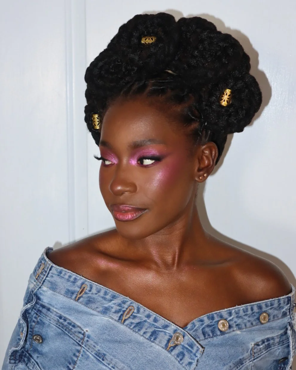 A woman with dark skin, styled in large, thick twist braids decorated with gold hair cuffs. She has pink eyeshadow, blush, and glossy lips, and is wearing an off-the-shoulder denim top.