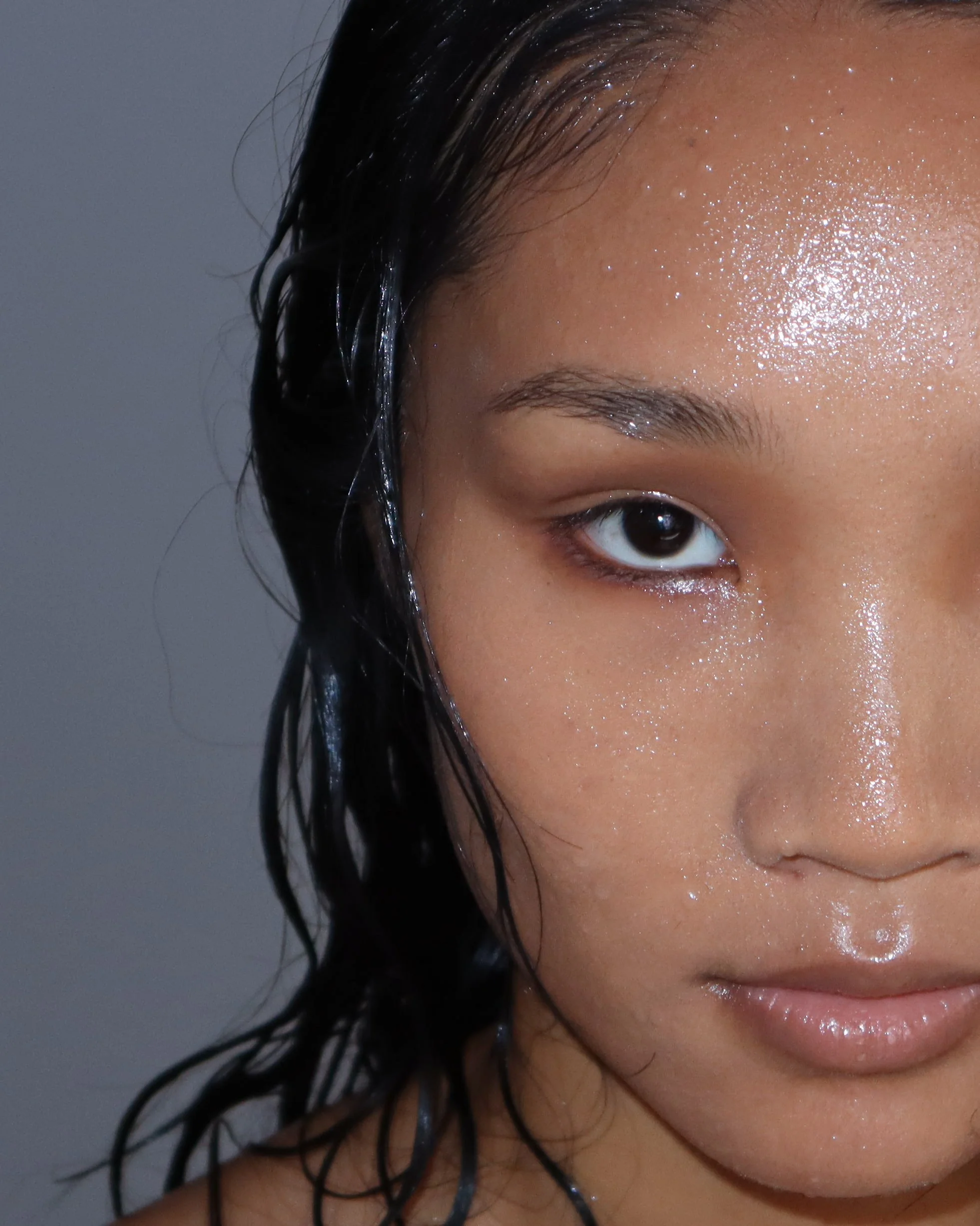 A close-up photo of a person's face showing the eye, eyebrow, part of the forehead, and wet hair against a plain grey background.