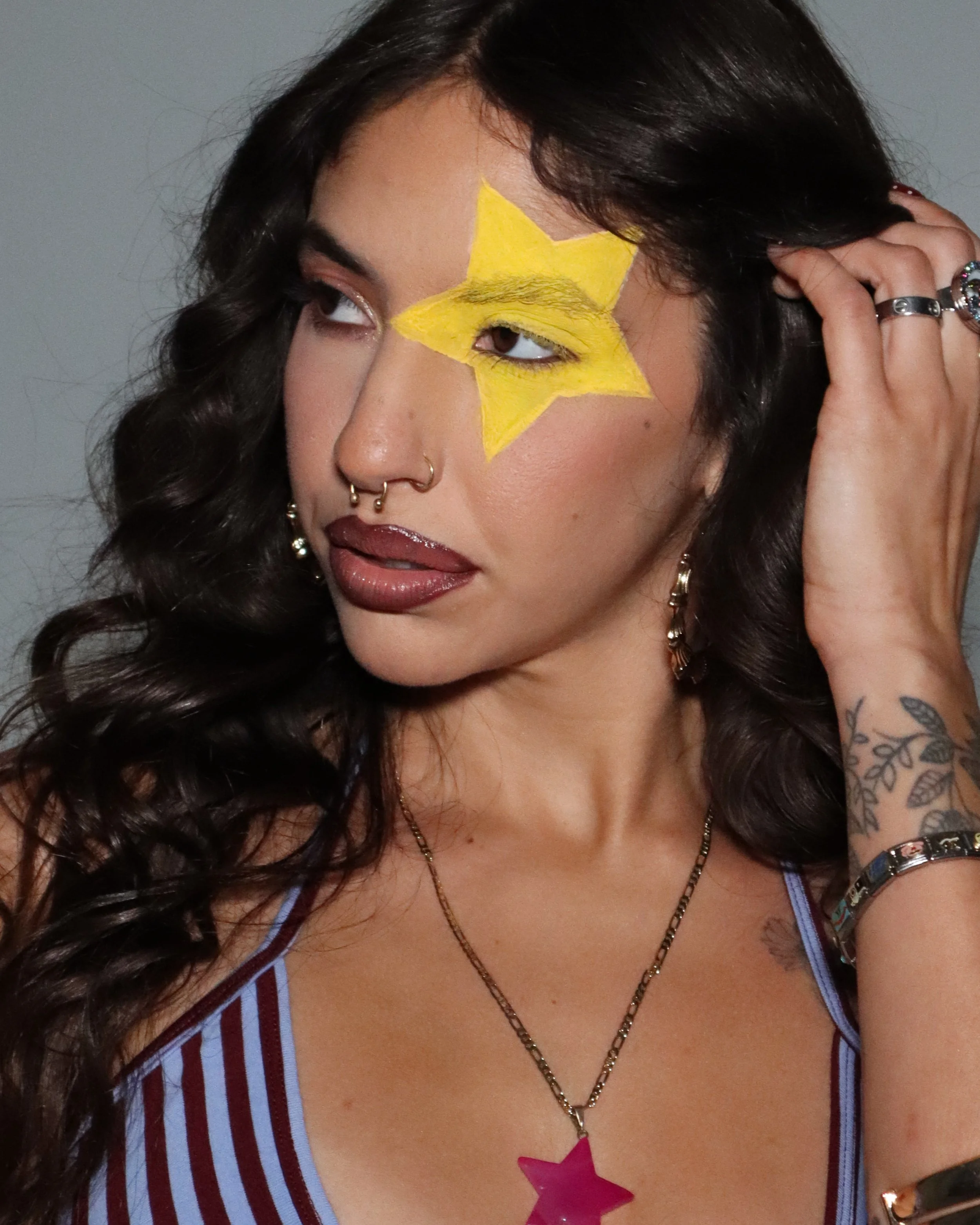 A young woman with dark, curly hair and tattoos on her arm, wearing necklaces, earrings, rings, and a striped top. She has a yellow star painted on her face, covering her eye.