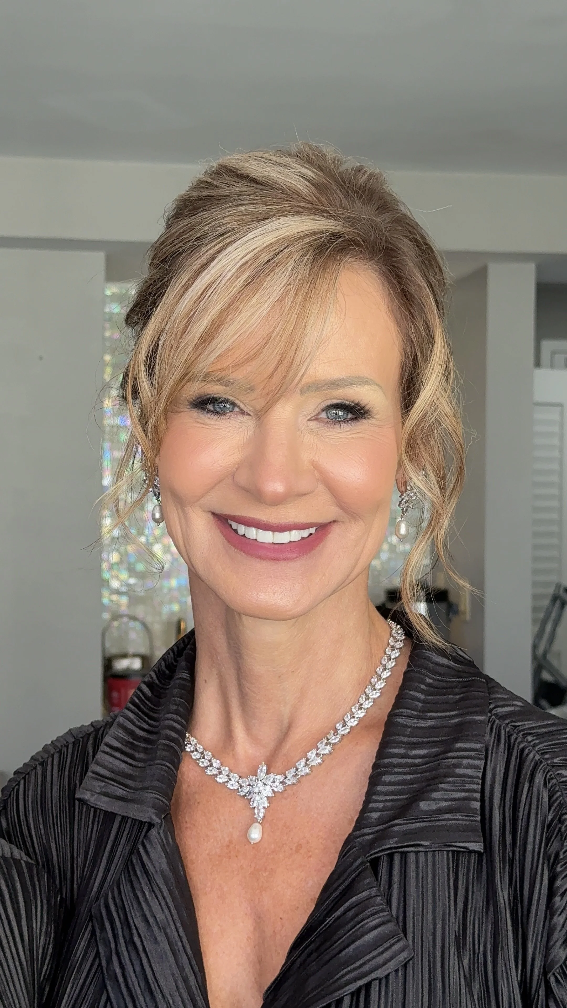 A smiling woman with short, styled blonde hair, wearing a black outfit and sparkling jewelry, in an indoor setting.
