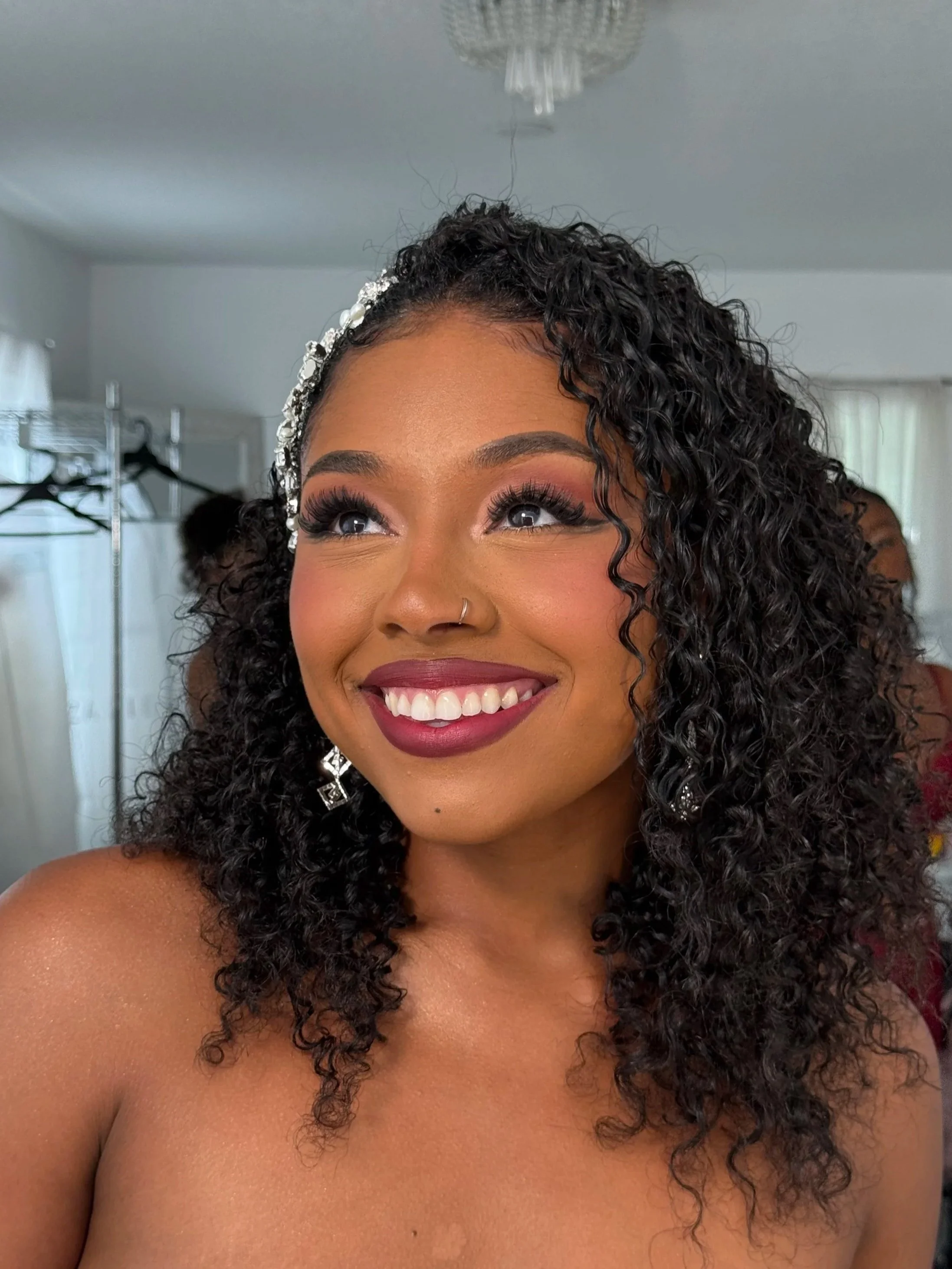 A woman with curly black hair styled with a pearl and rhinestone headband, smiling, showing white teeth and wearing dark lipstick, in a room with a closet and other people in the background.