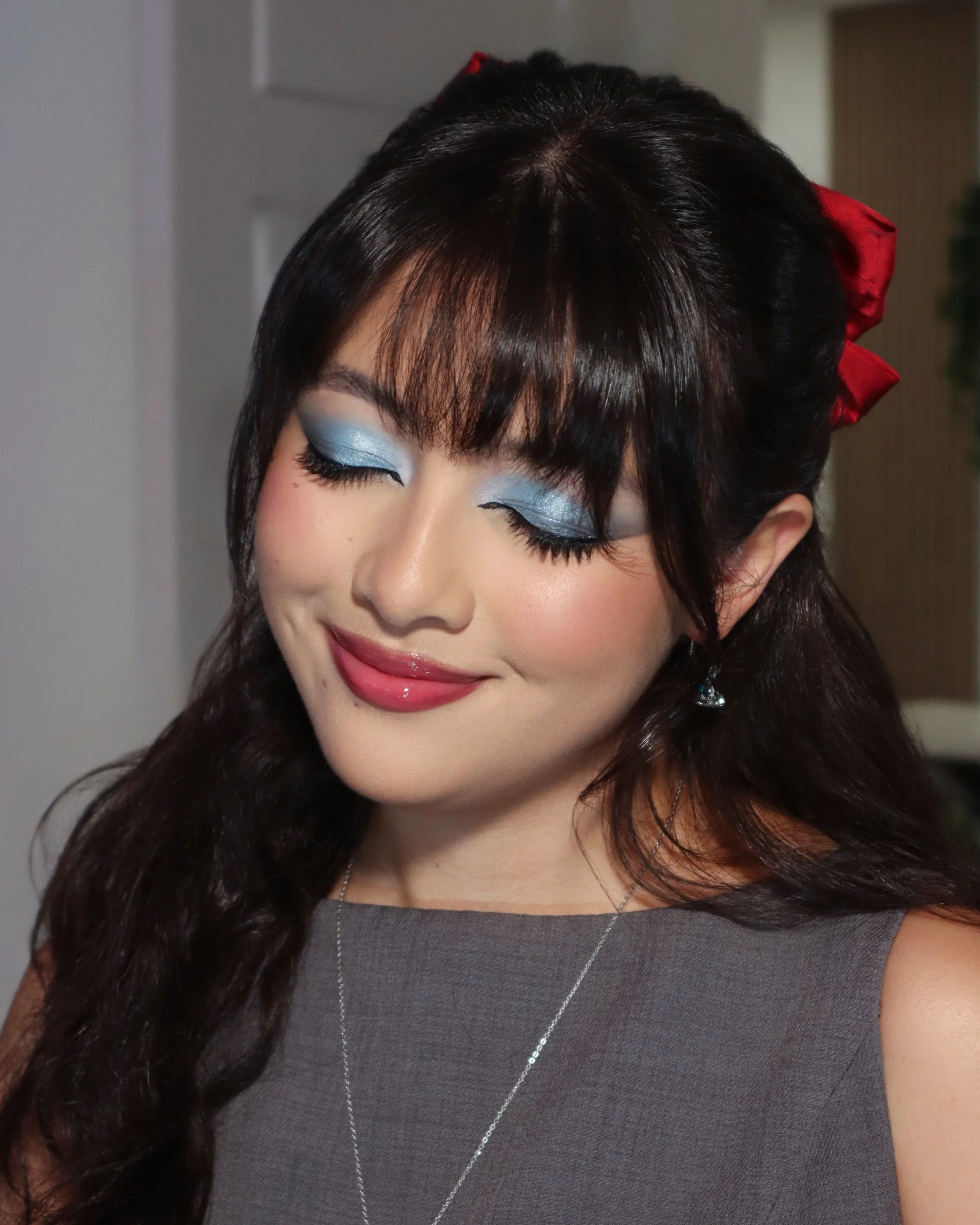 A woman with long dark hair styled with waves and a red bow, wearing eye makeup with blue eyeshadow, lipstick, earrings, and a silver necklace. She is smiling with closed eyes.