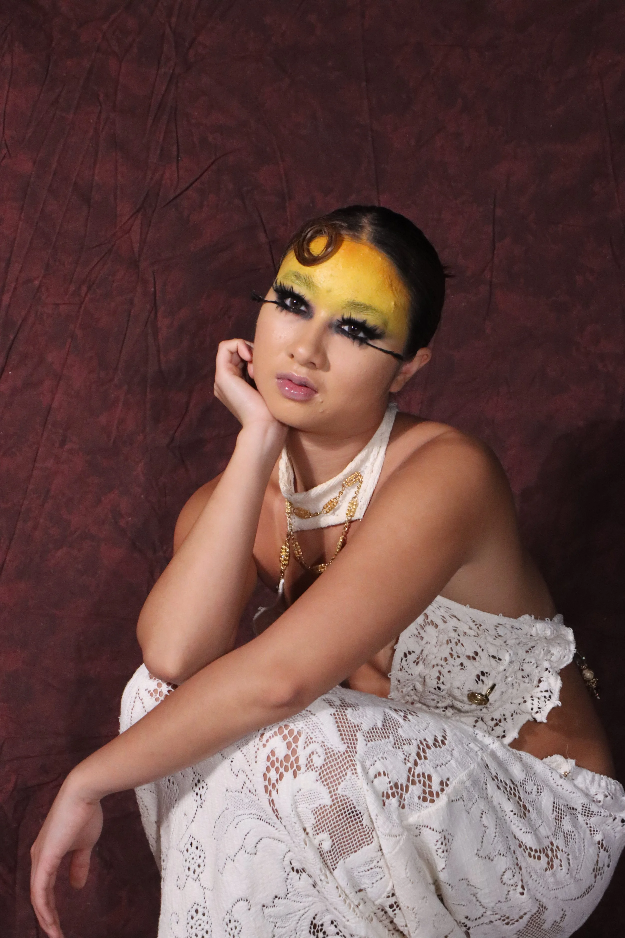 A person with makeup, including bright yellow face paint, long false eyelashes, and dramatic eye makeup, poses against a textured dark red background. They are wearing a white lace outfit and jewelry.