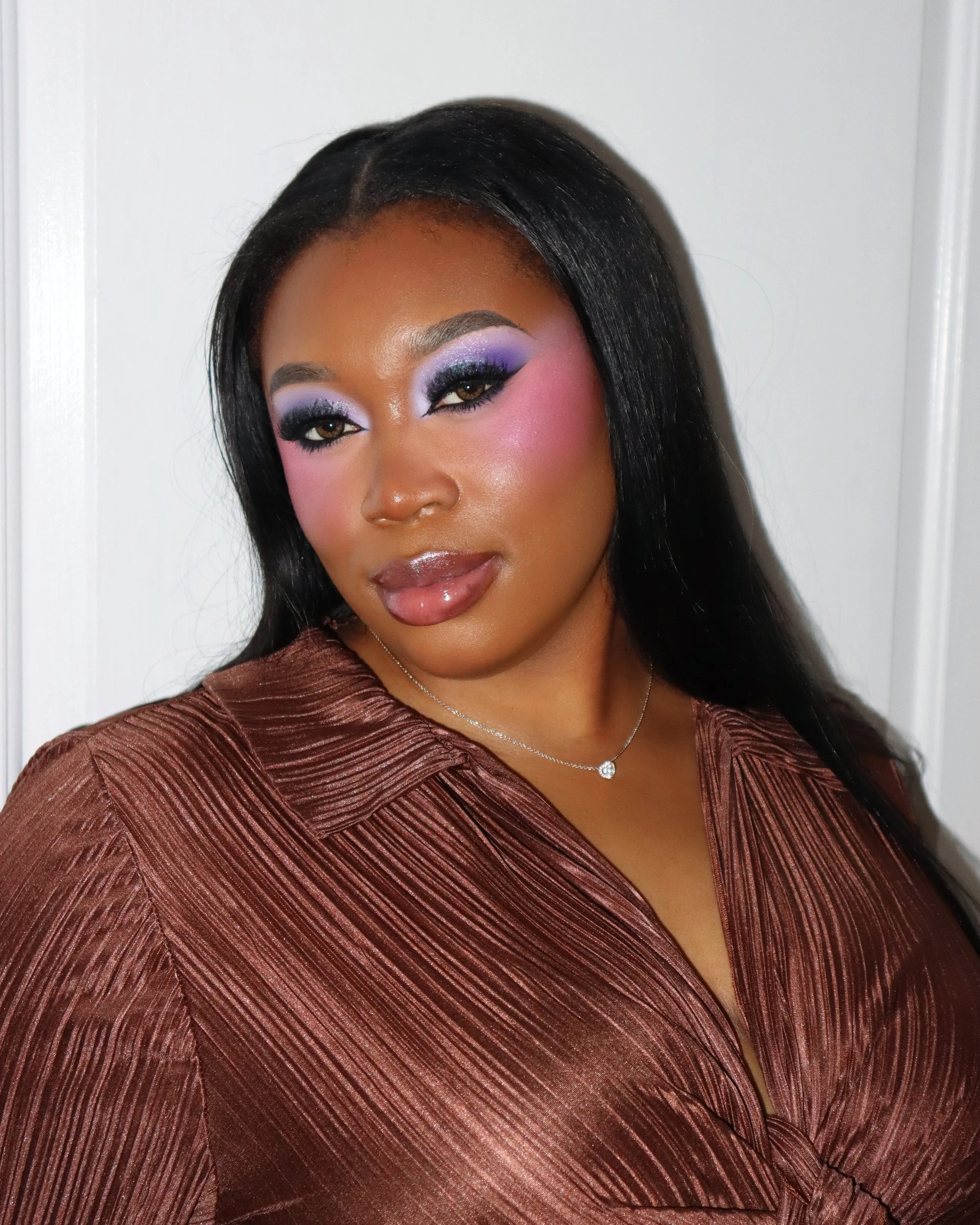 A woman with long black hair, wearing makeup with purple eyeshadow, pink blush, and glossy lips, dressed in a shiny brown top, standing against a white door.