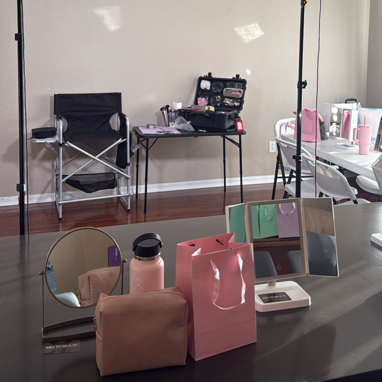 A makeup or beauty station set up indoors with mirrors, pink gift bags, a water bottle, and makeup tools on a table, with a backdrop of chairs and equipment likely for a photo shoot or event.
