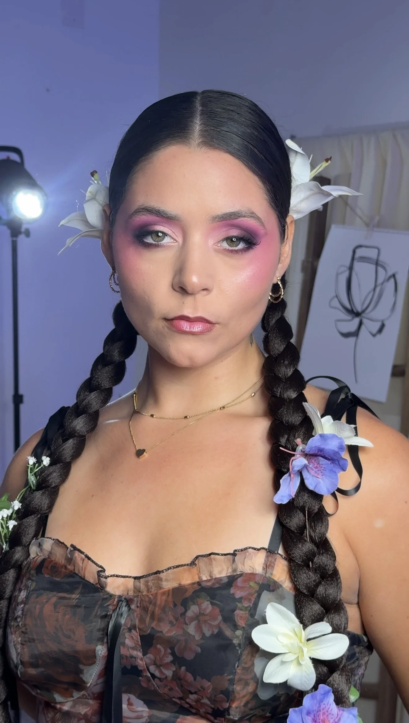 A woman with long braided hair decorated with white and purple flowers, wearing pink eyeshadow, eyeliner, and a floral dress, standing in a studio with a backdrop and bright lights.